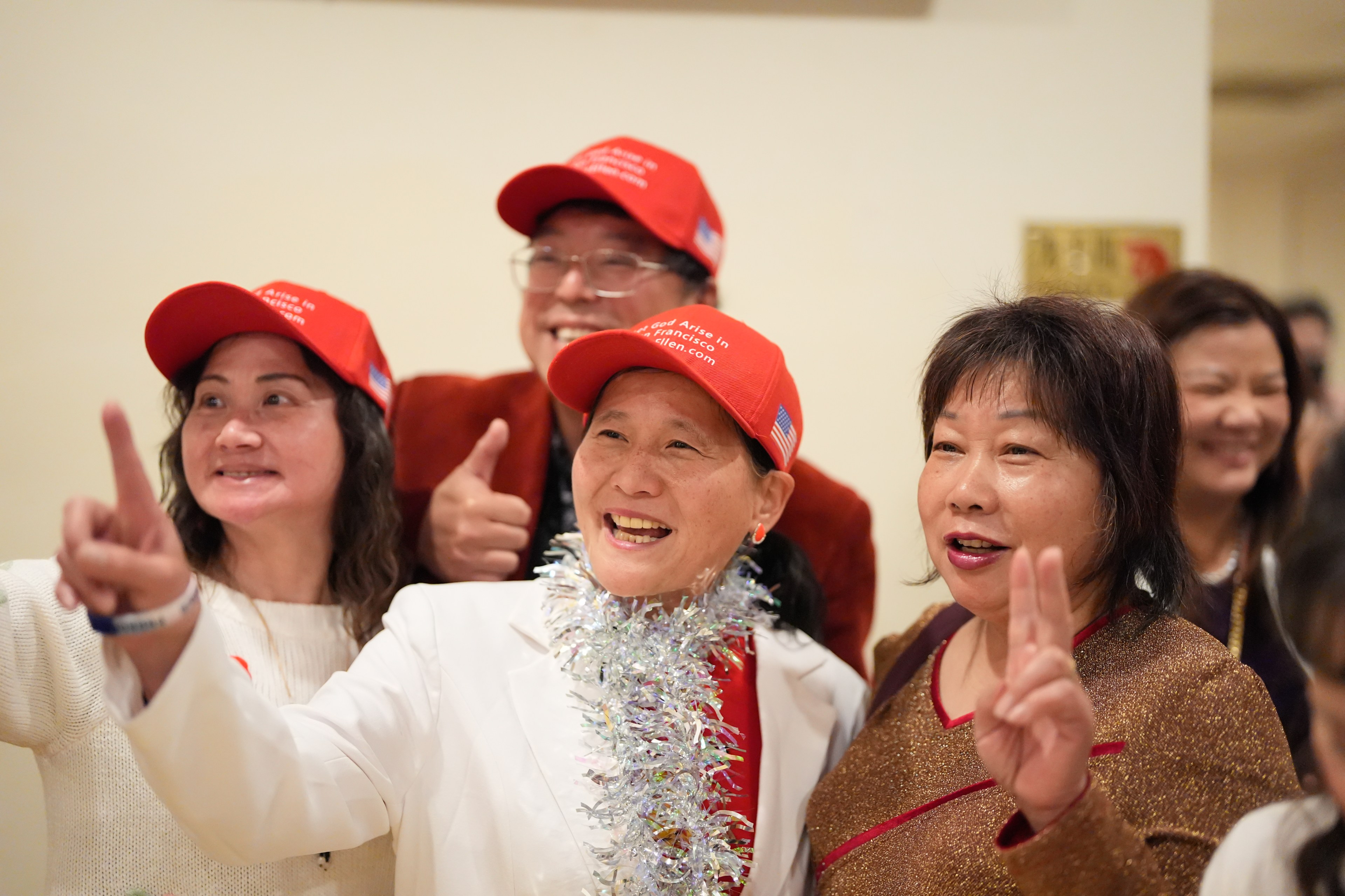 A group of joyous people, some wearing red caps, is gesturing with thumbs up and peace signs.