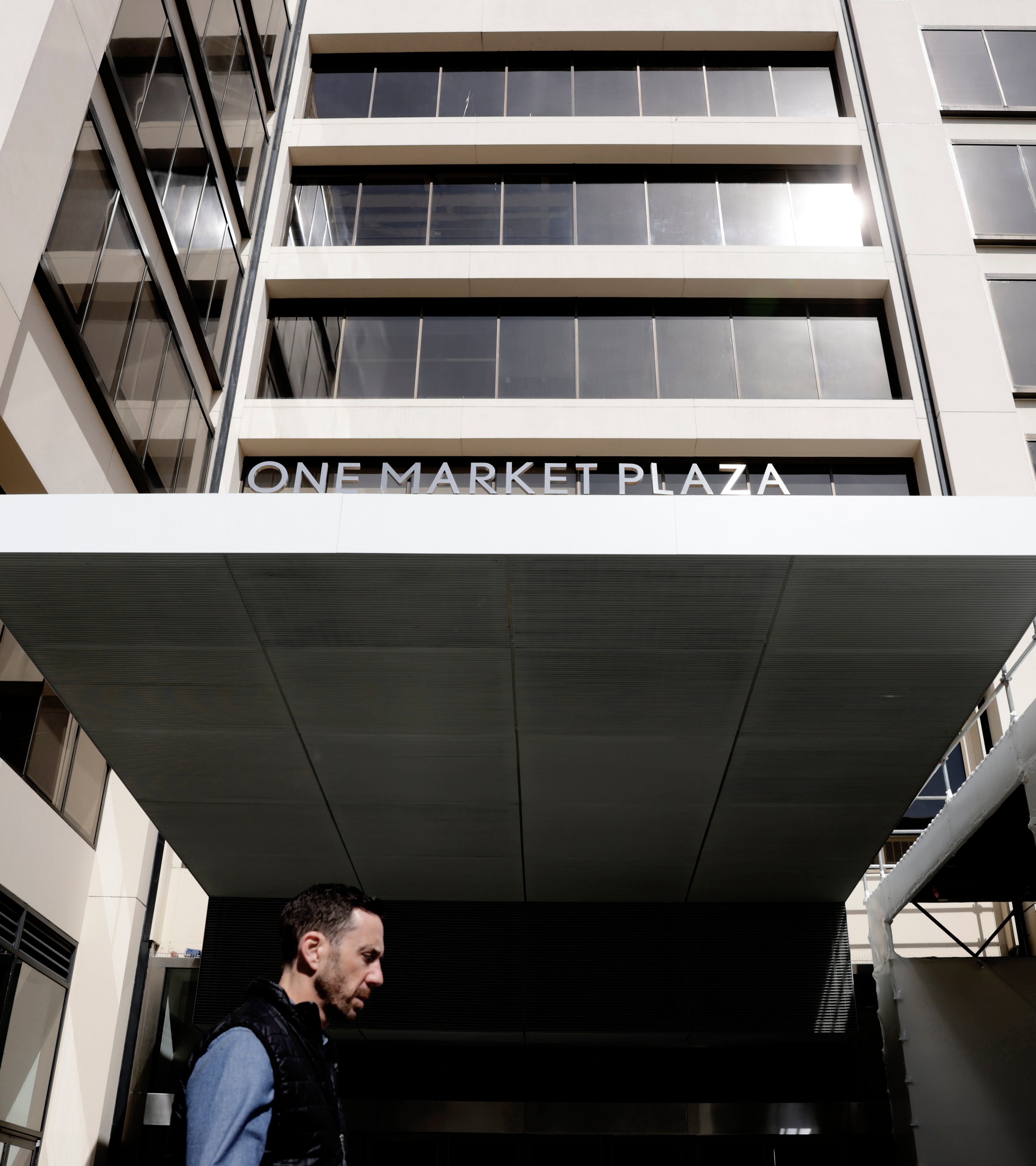 A building labeled &quot;One Market Plaza&quot; with a man in a black vest and blue shirt walking in front. The sun reflects off the windows above.