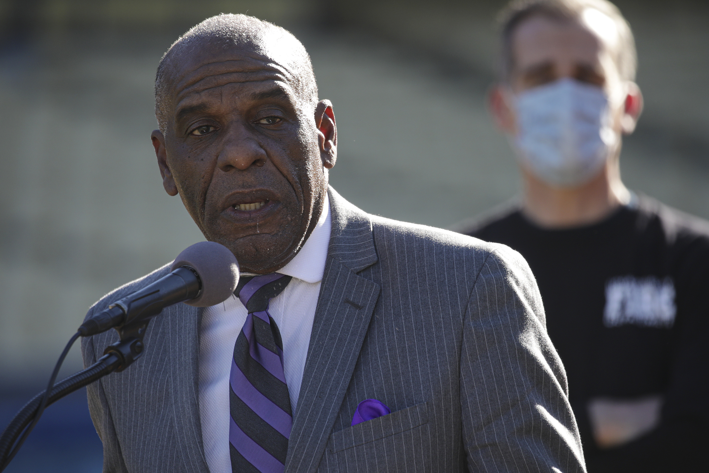An older man in a suit speaks at a microphone; another man wearing a mask stands out of focus in the background.