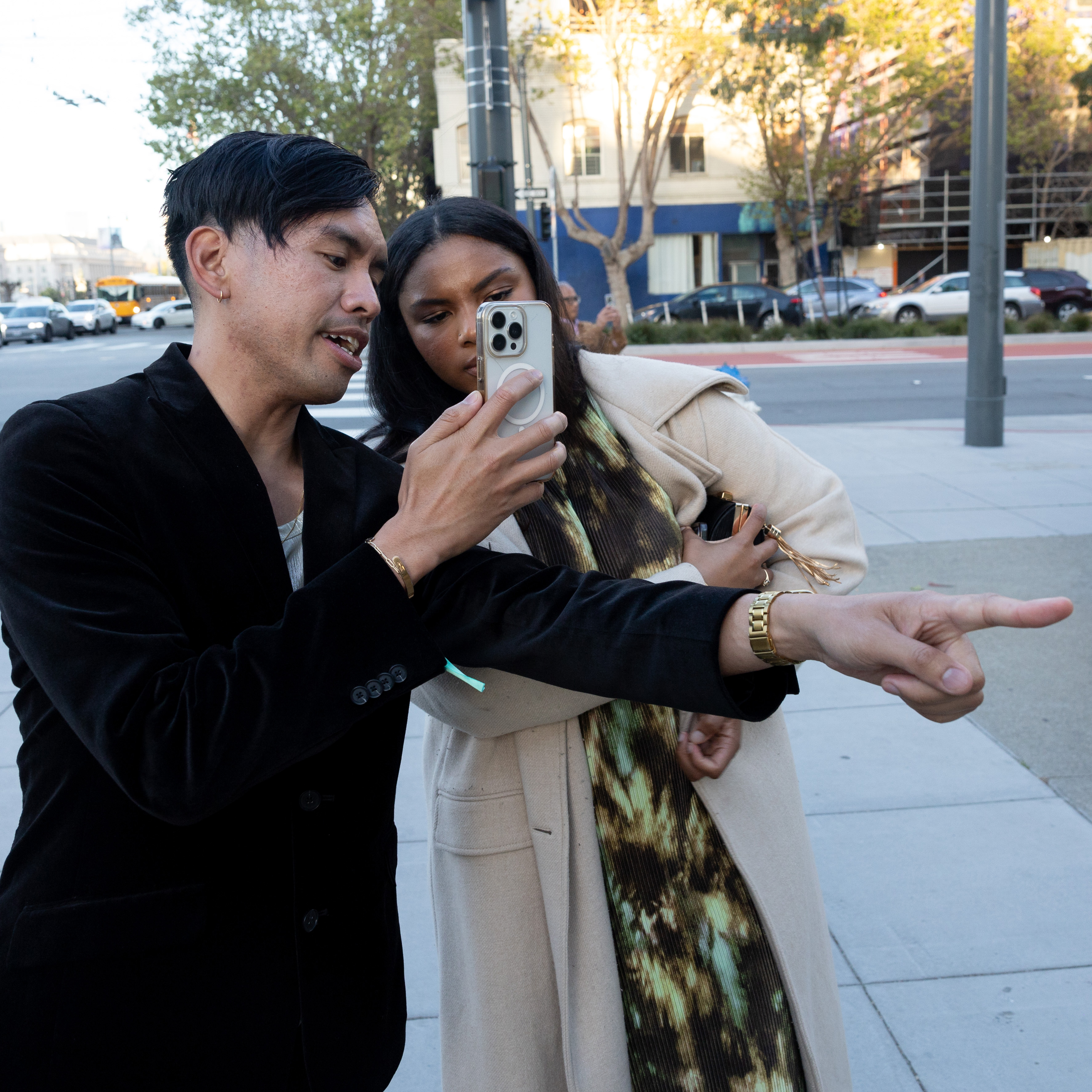 Two people are posing for a selfie; one points away, the other holds the phone, with a city street in the background.
