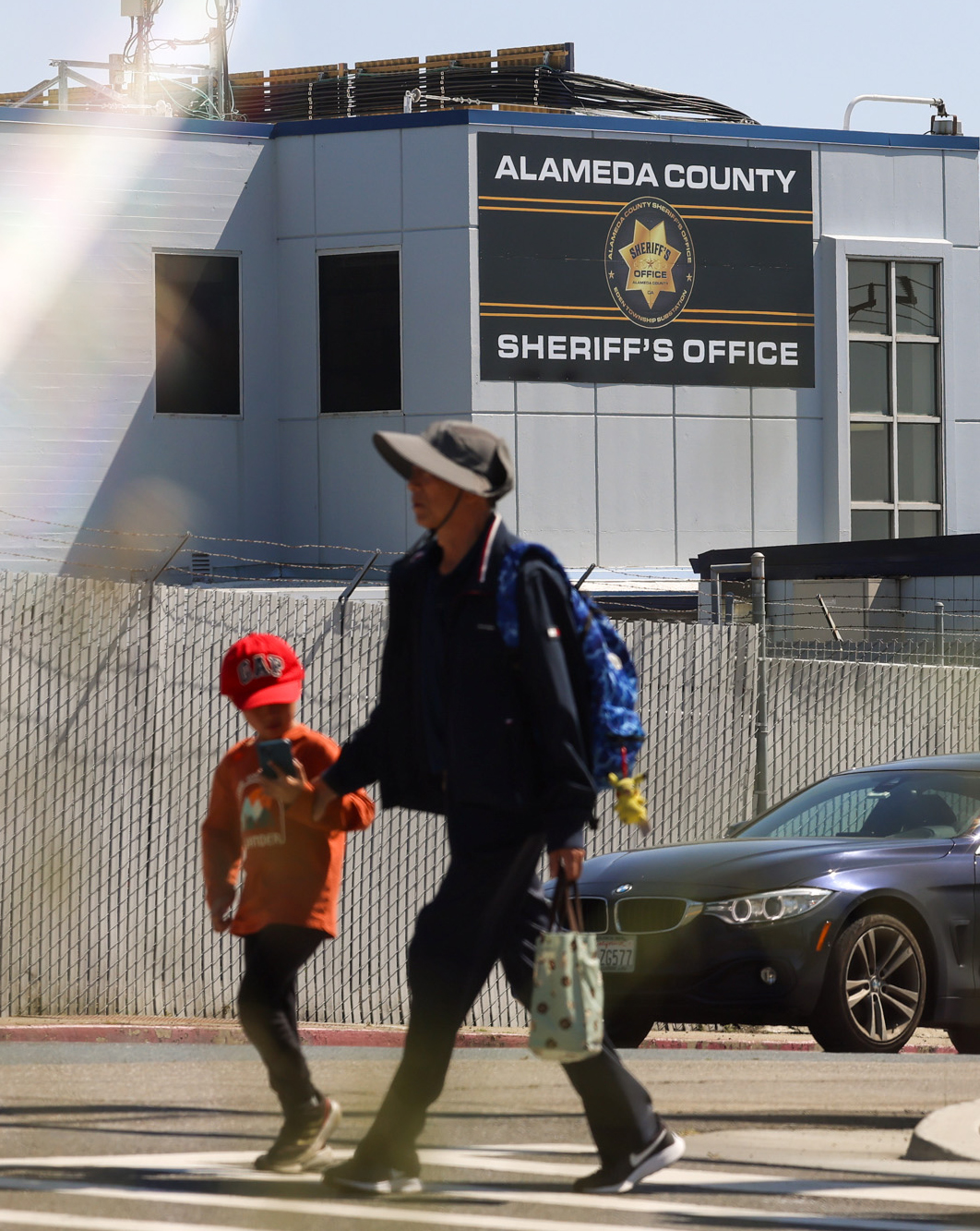 An adult and child walk by a building labeled &quot;ALAMEDA COUNTY SHERIFF'S OFFICE&quot; with a car passing in the background.