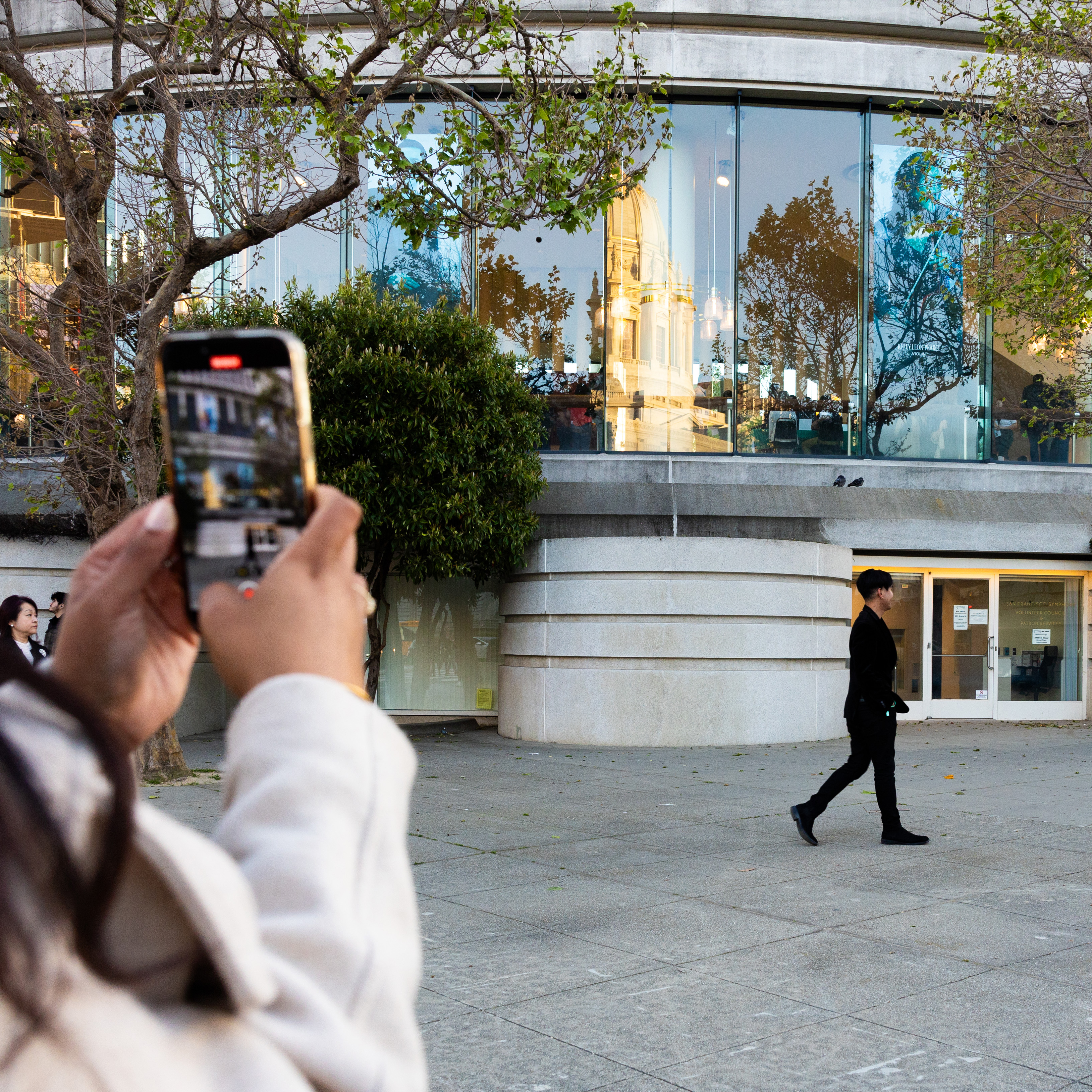 A person is taking a photo with their phone capturing another person walking by a building with reflective windows.
