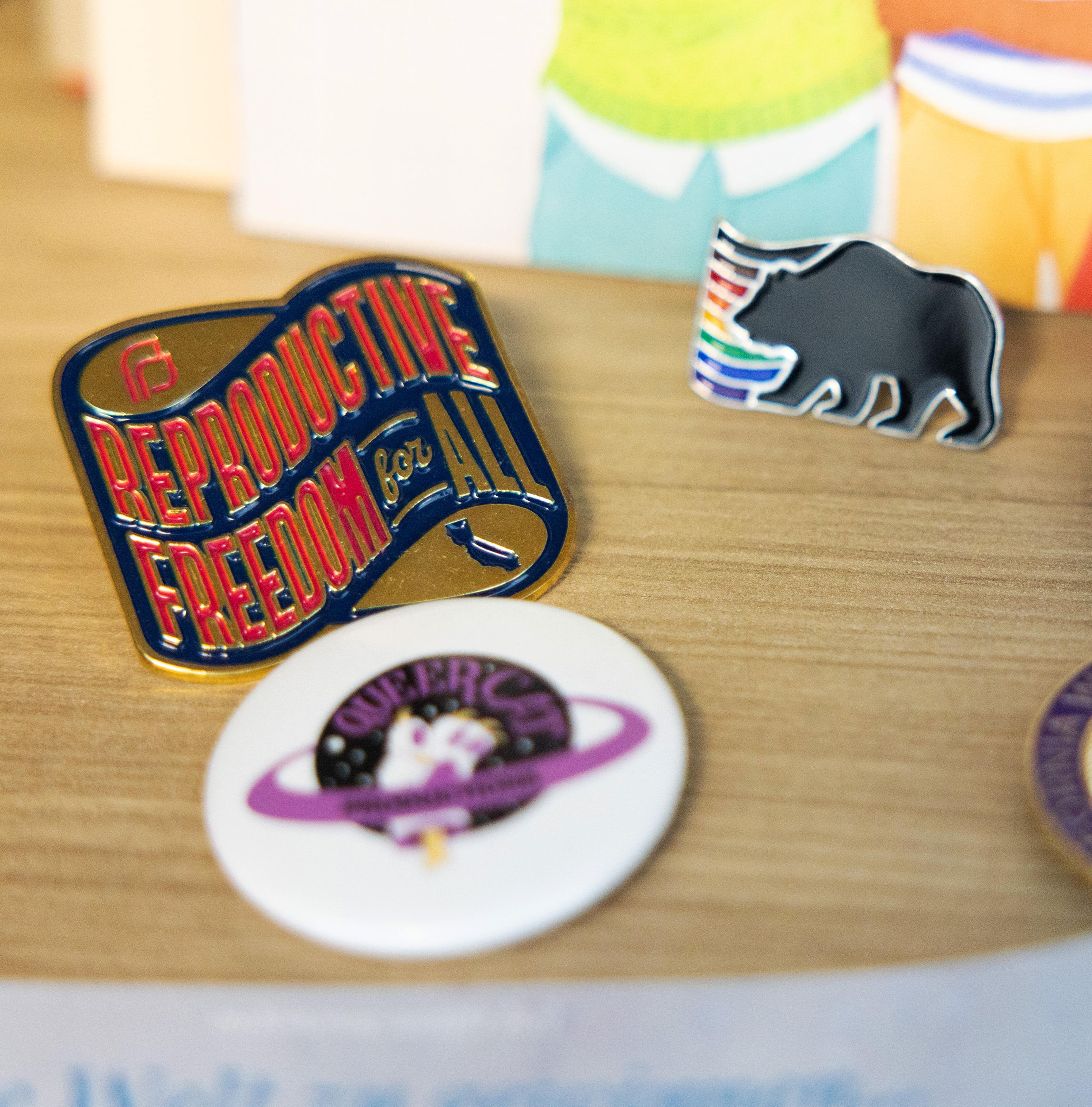 Enamel pins with messages like &quot;Reproductive Freedom For All&quot; &amp; graphics, like a bear with colorful stripes, on a wood surface.