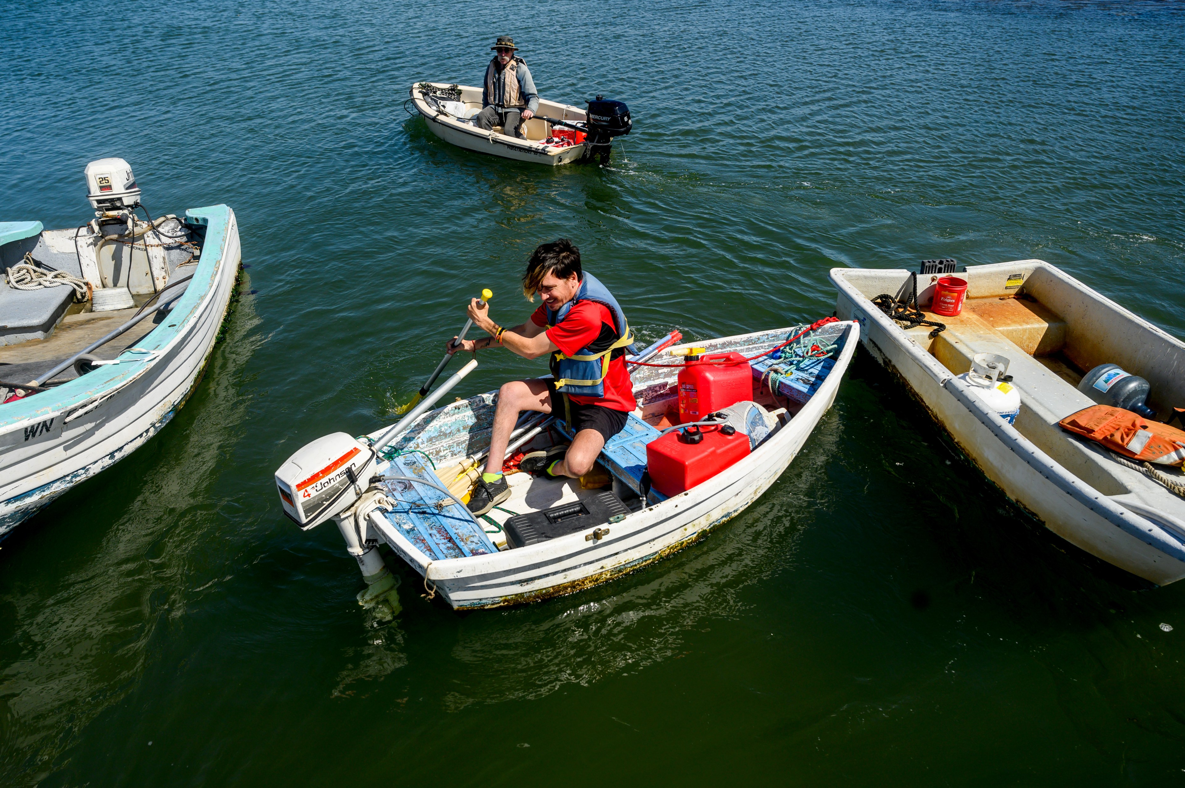 A person in a red life jacket rows a cluttered small boat among others on a sunny, calm water body.