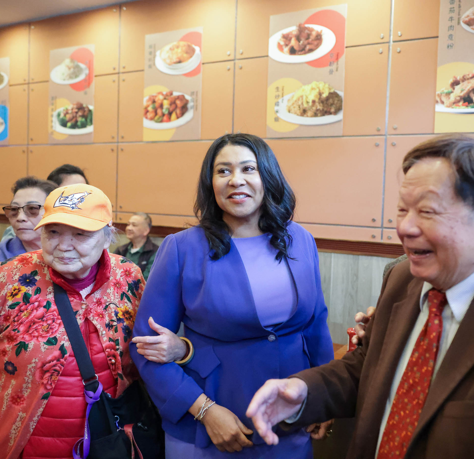 A woman in a blue outfit smiles while shaking hands with a man in a restaurant with meal photos on the wall; an elderly lady in a floral jacket observes.