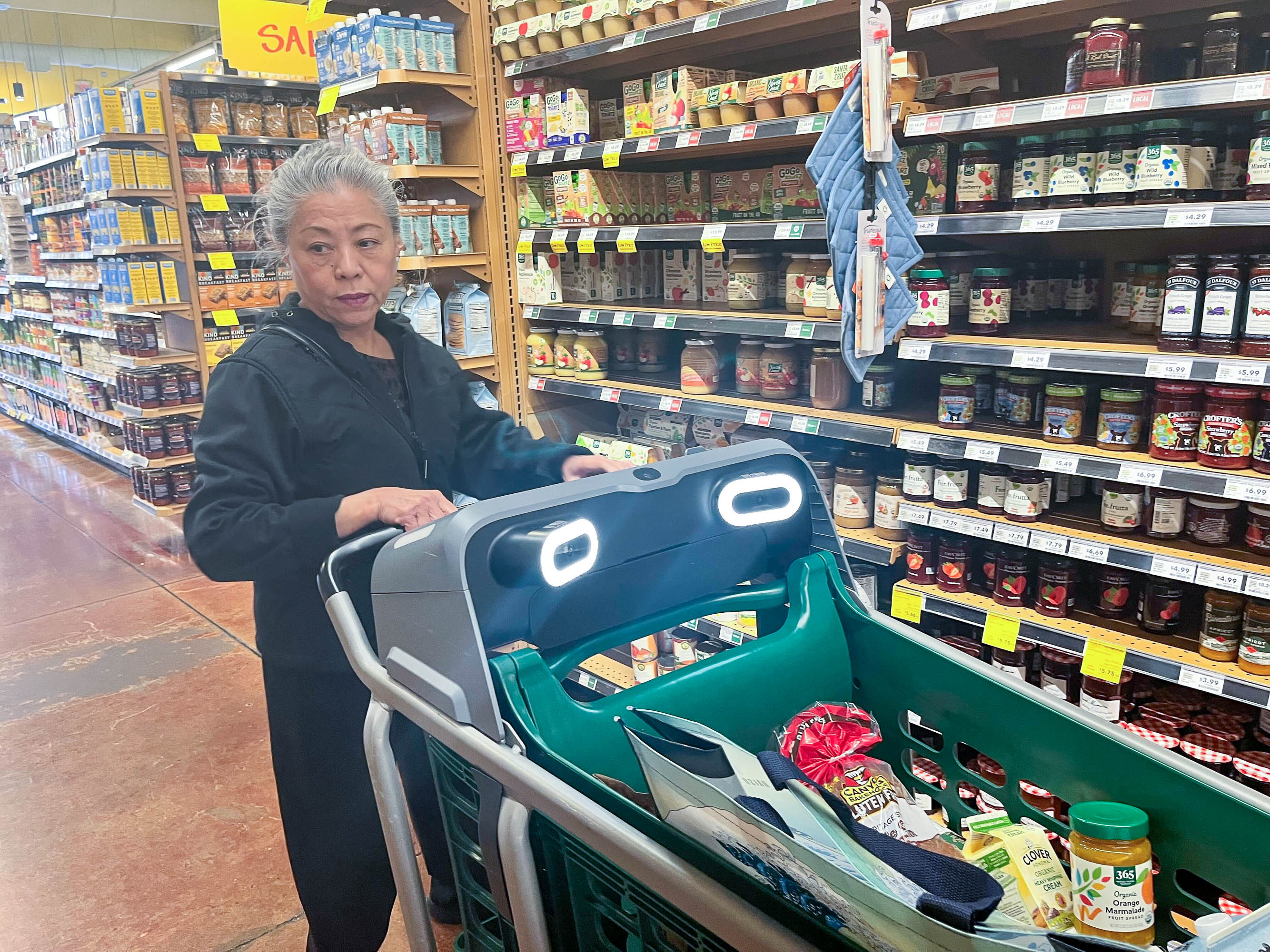 A woman with gray hair shops in a grocery aisle, pushing a cart with items in it.