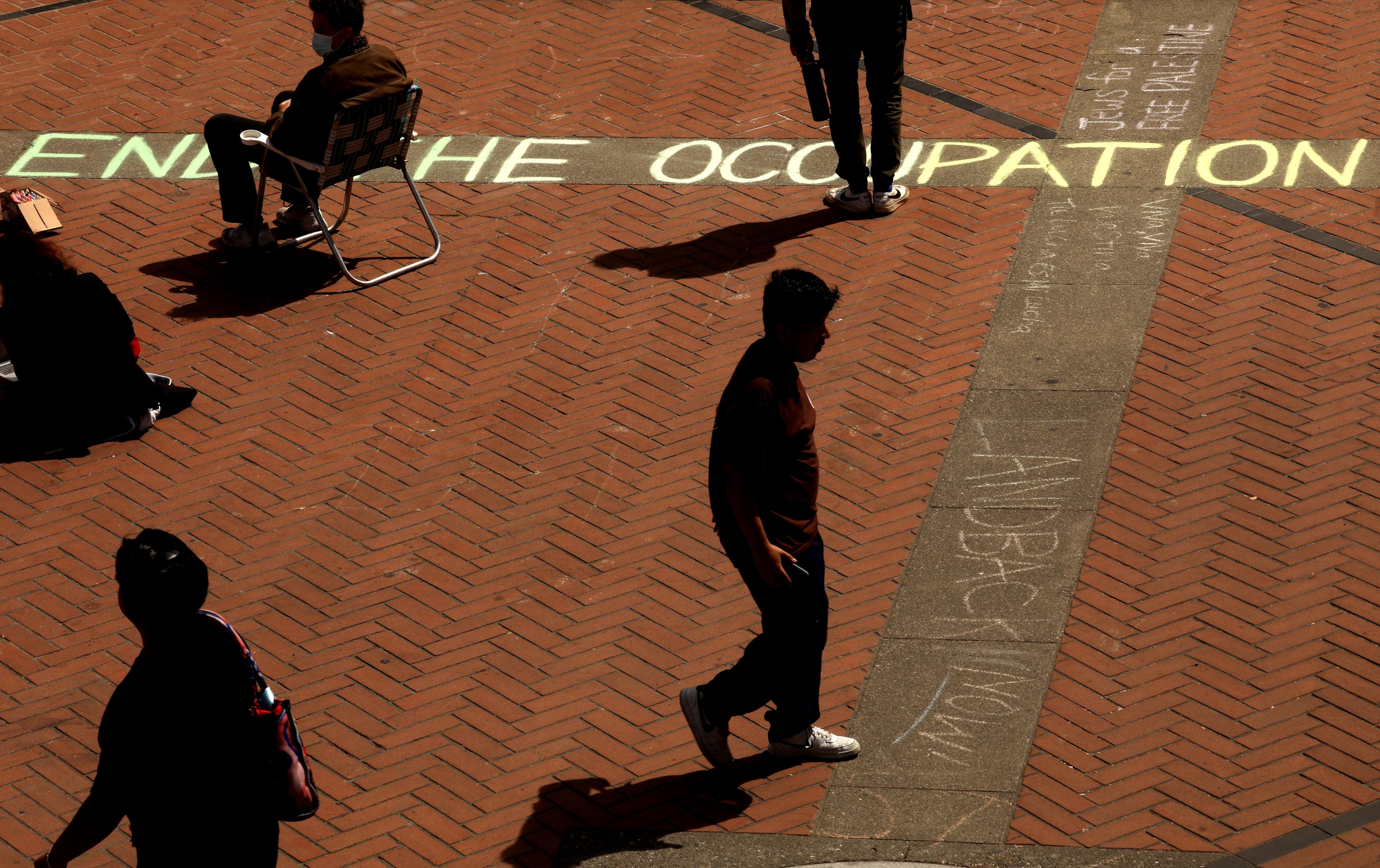 People walk over brick pavement with the phrase &quot;END THE OCCUPATION&quot; chalked in large letters. Shadows suggest bright sunlight.