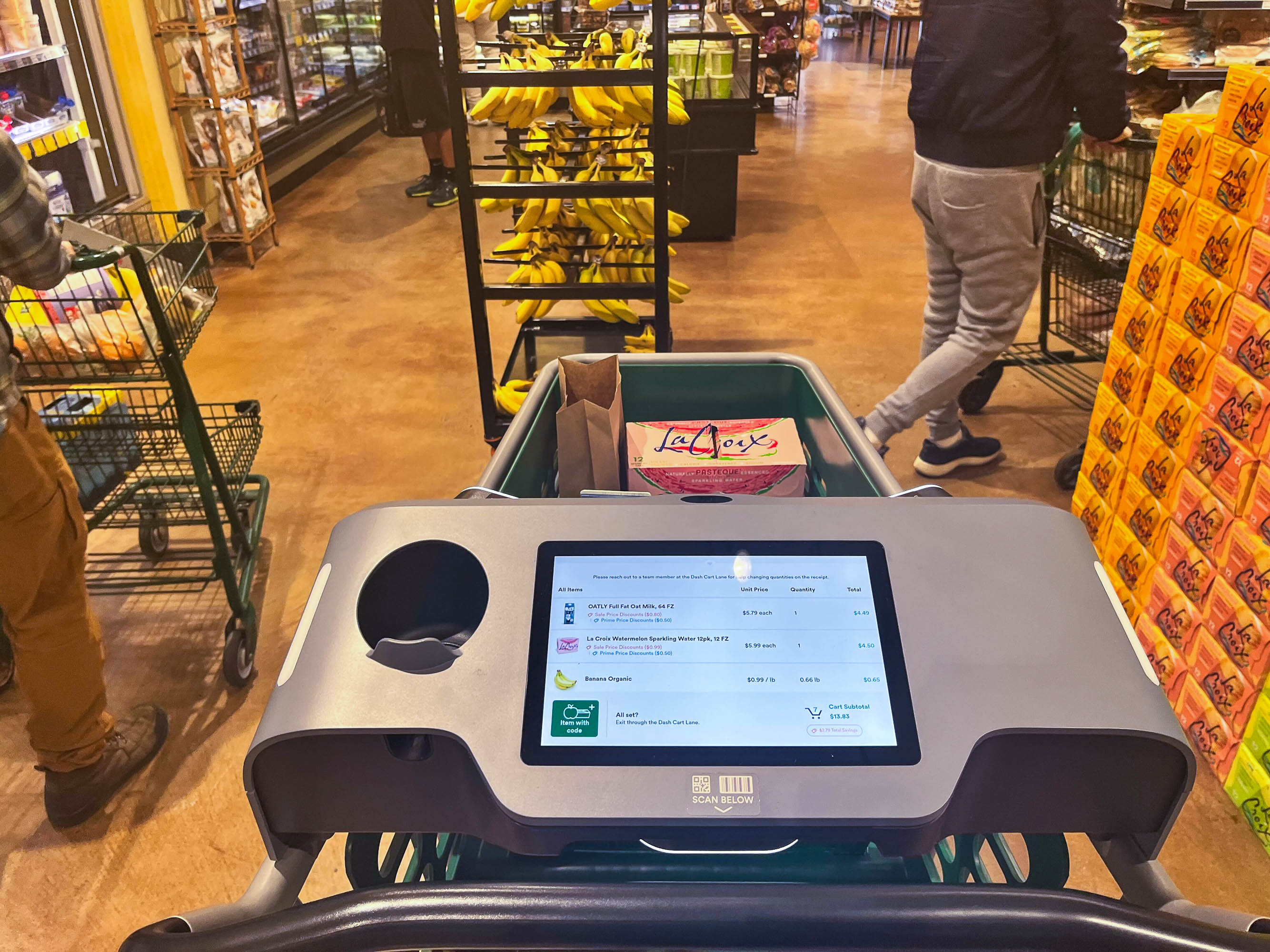 A shopping cart with a digital screen displaying items, in a grocery store aisle with shoppers and produce nearby.