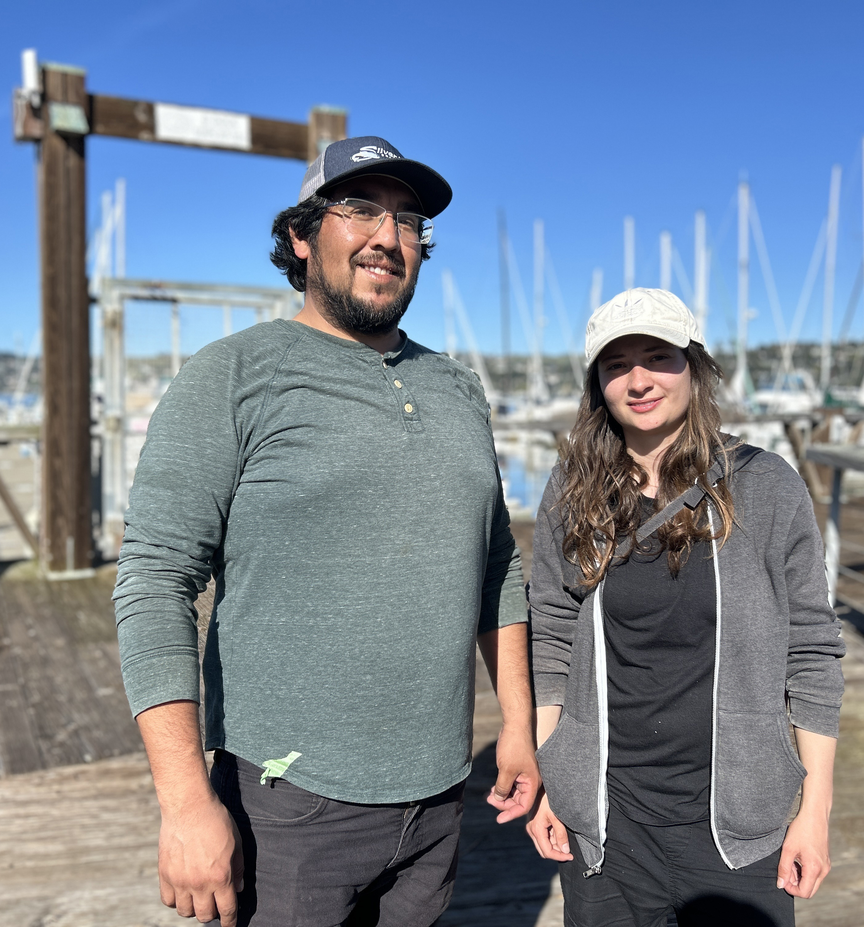 Two people are standing by a marina with boats and clear skies in the background. They are casually dressed and smiling.