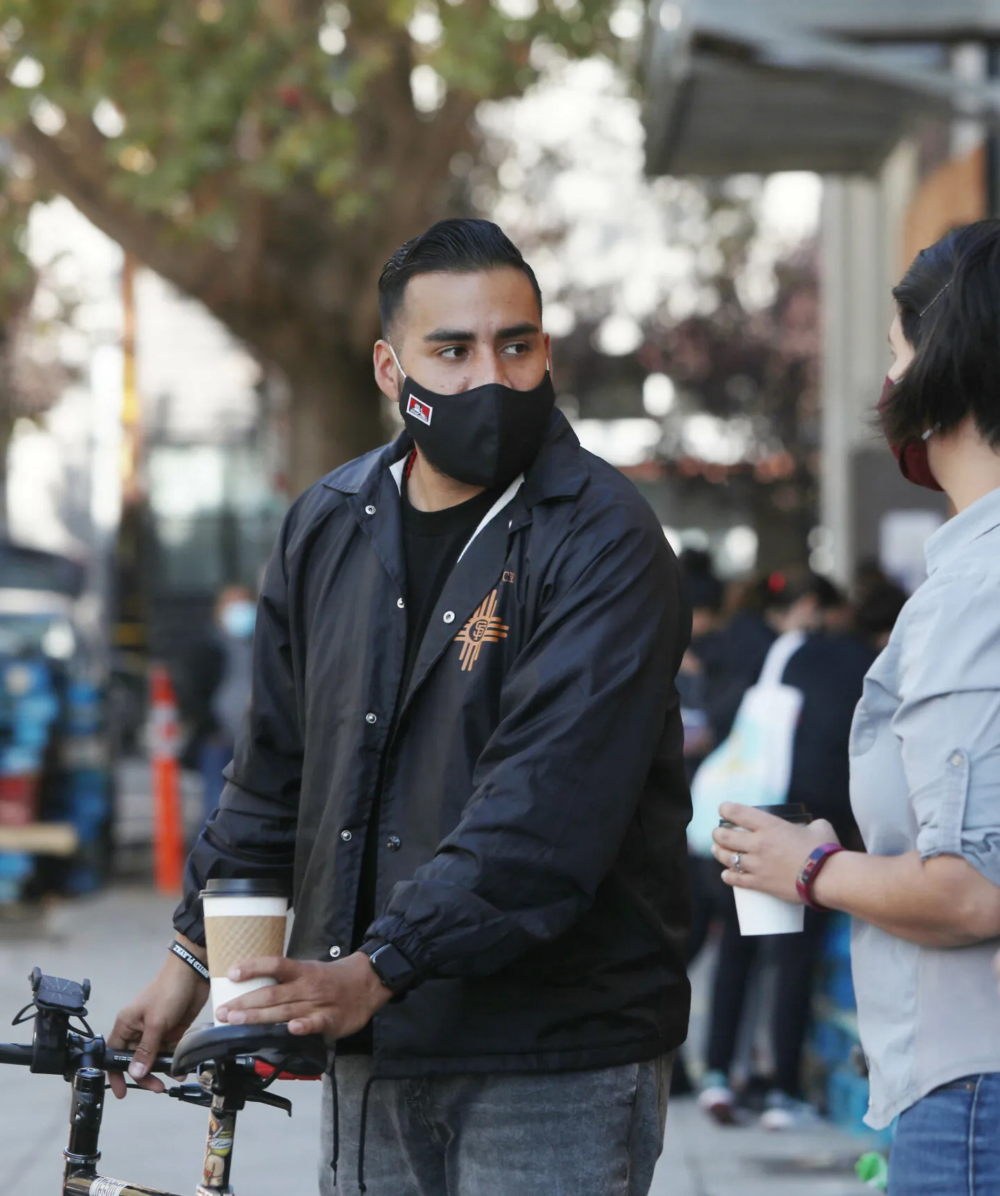 Jon Jacobo in a mask holding a coffee stands next to his bike, talking to an out-of-focus person.