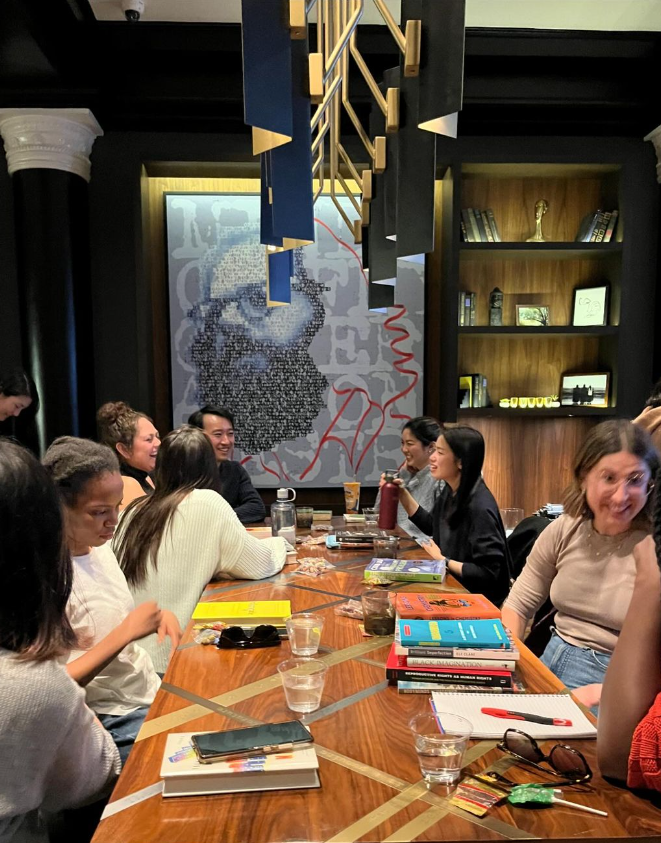 A group of people gathered around a table with books, engaged in conversation in an elegant room with a striking art piece on the wall.