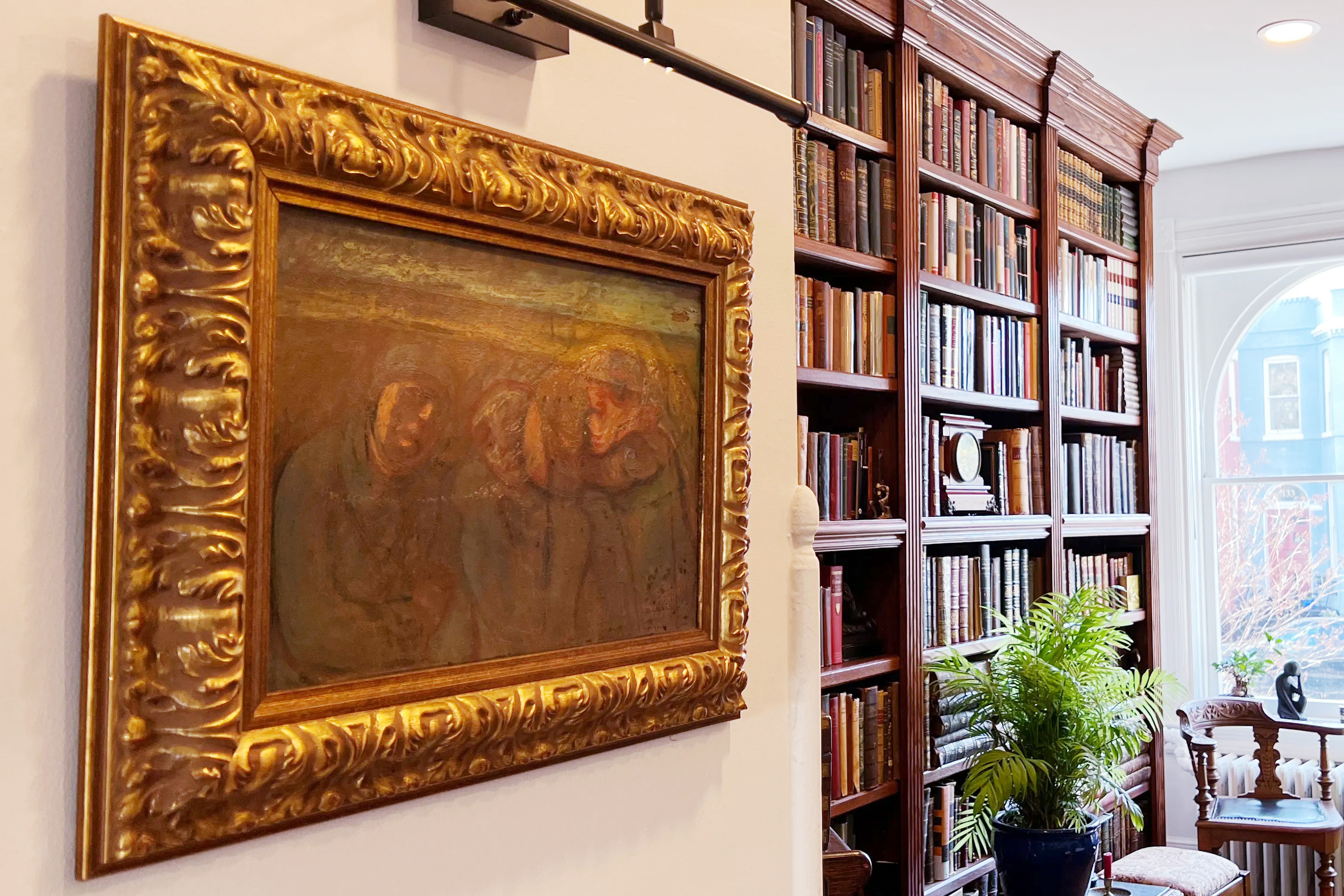 A classic painting in a gilded frame beside a bookshelf-lined wall in a cozy room with a plant and window.