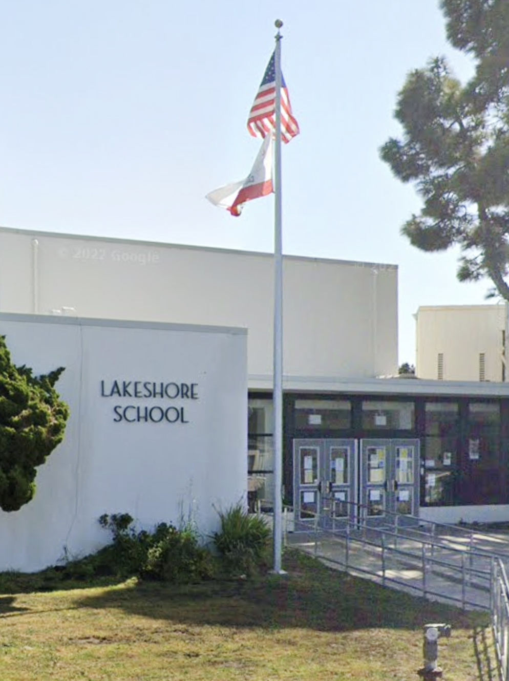 A flagpole with US &amp; California flags flies before &quot;Lakeshore School&quot; with glass doors and a ramp.