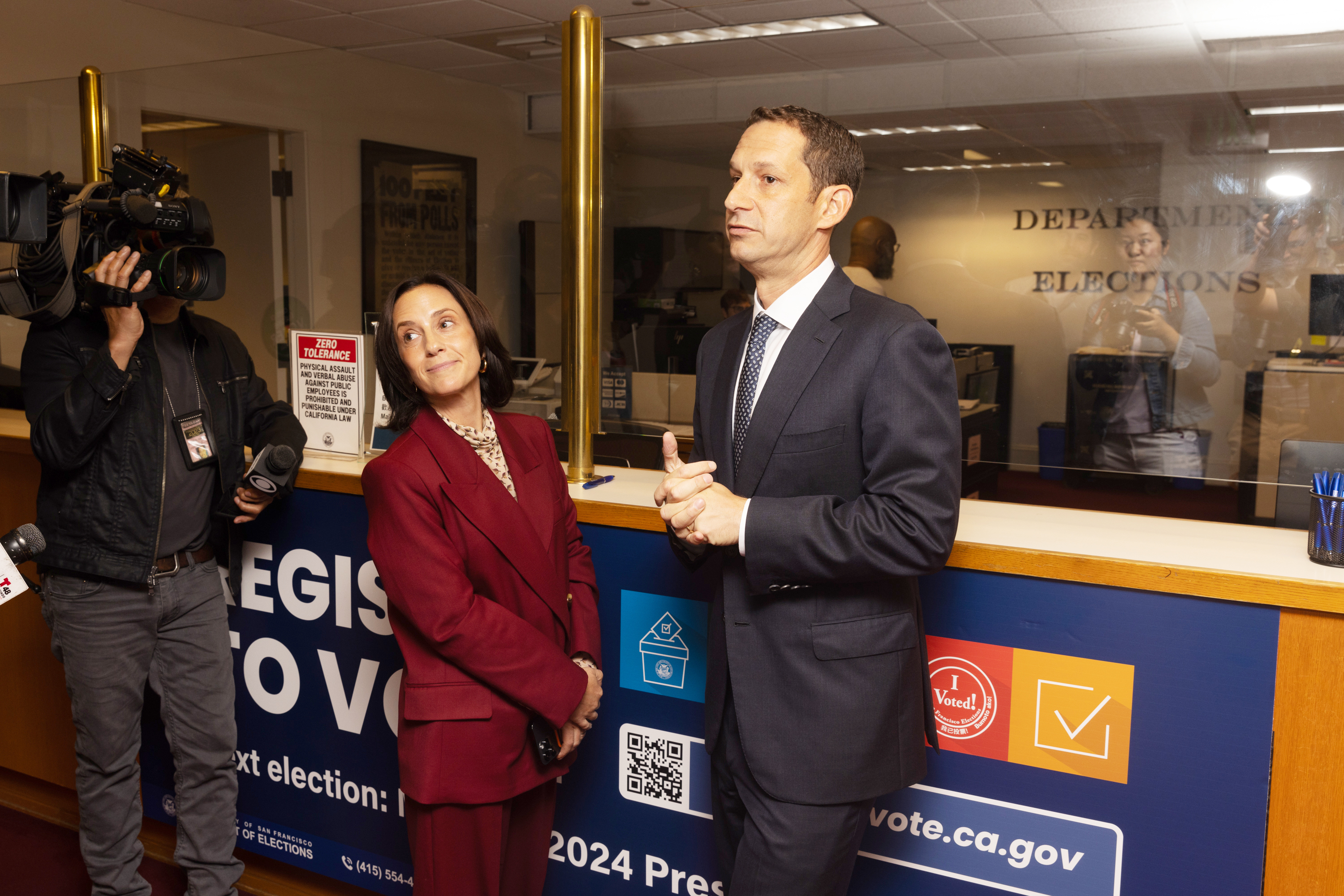 Becca Prowda and Daniel Lurie stand at a counter with &quot;Department of Elections&quot; signage.