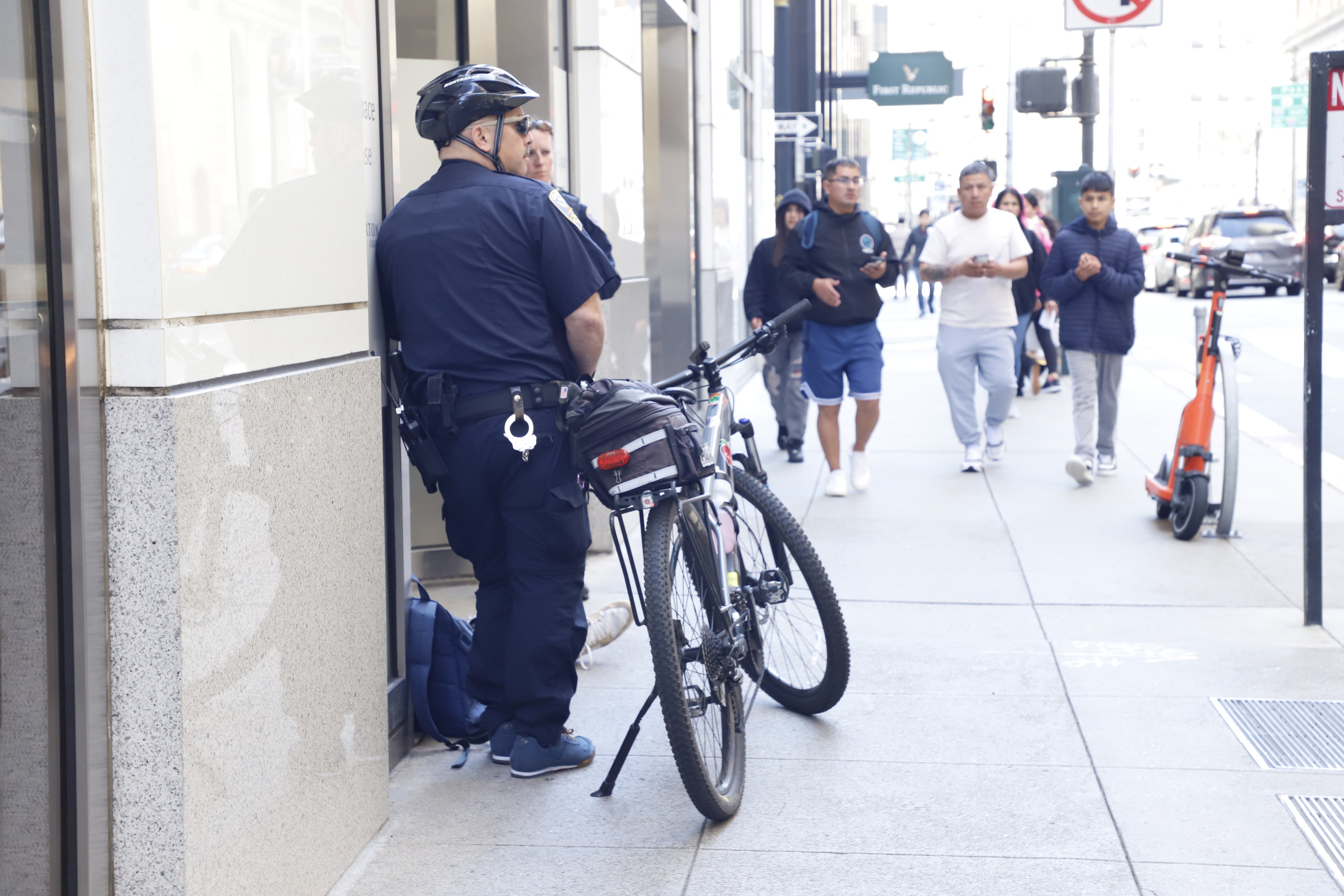 Two police officers are standing by a bike on a sunny city sidewalk as people walk by.