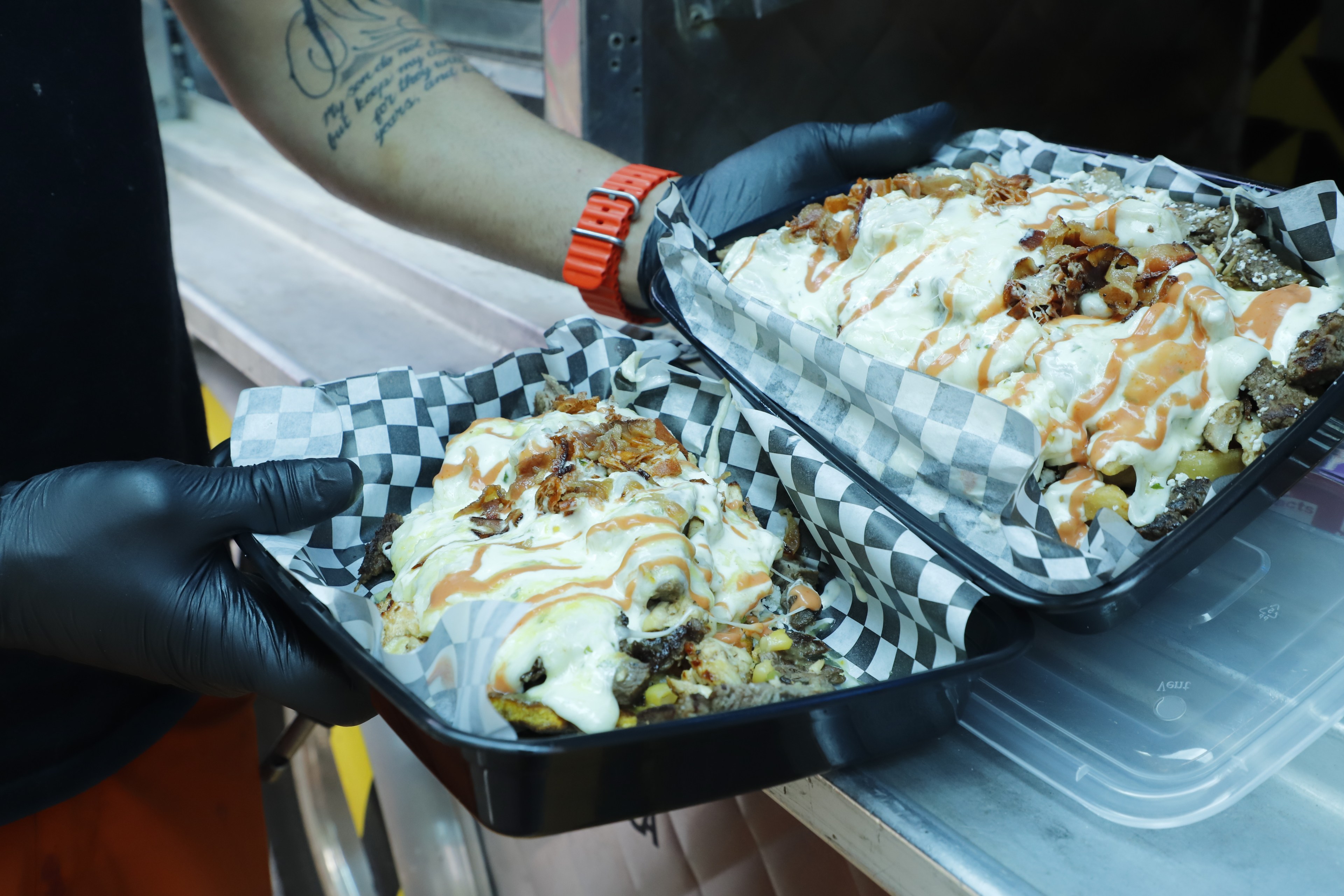 A person in gloves holds a tray of loaded fries with various toppings and sauces.