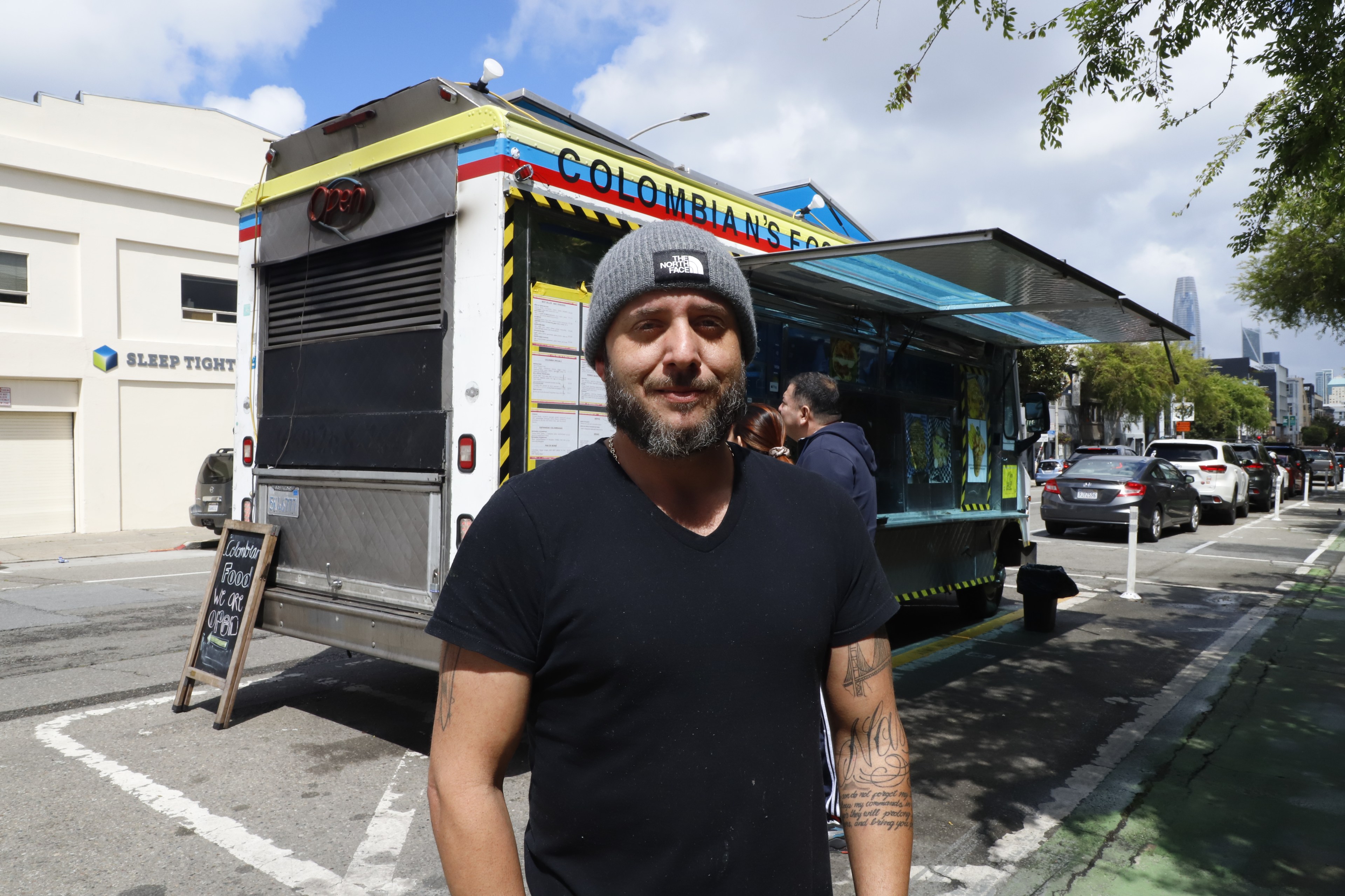 A man in a beanie stands smiling in front of a colorful Colombian food truck on a city street.
