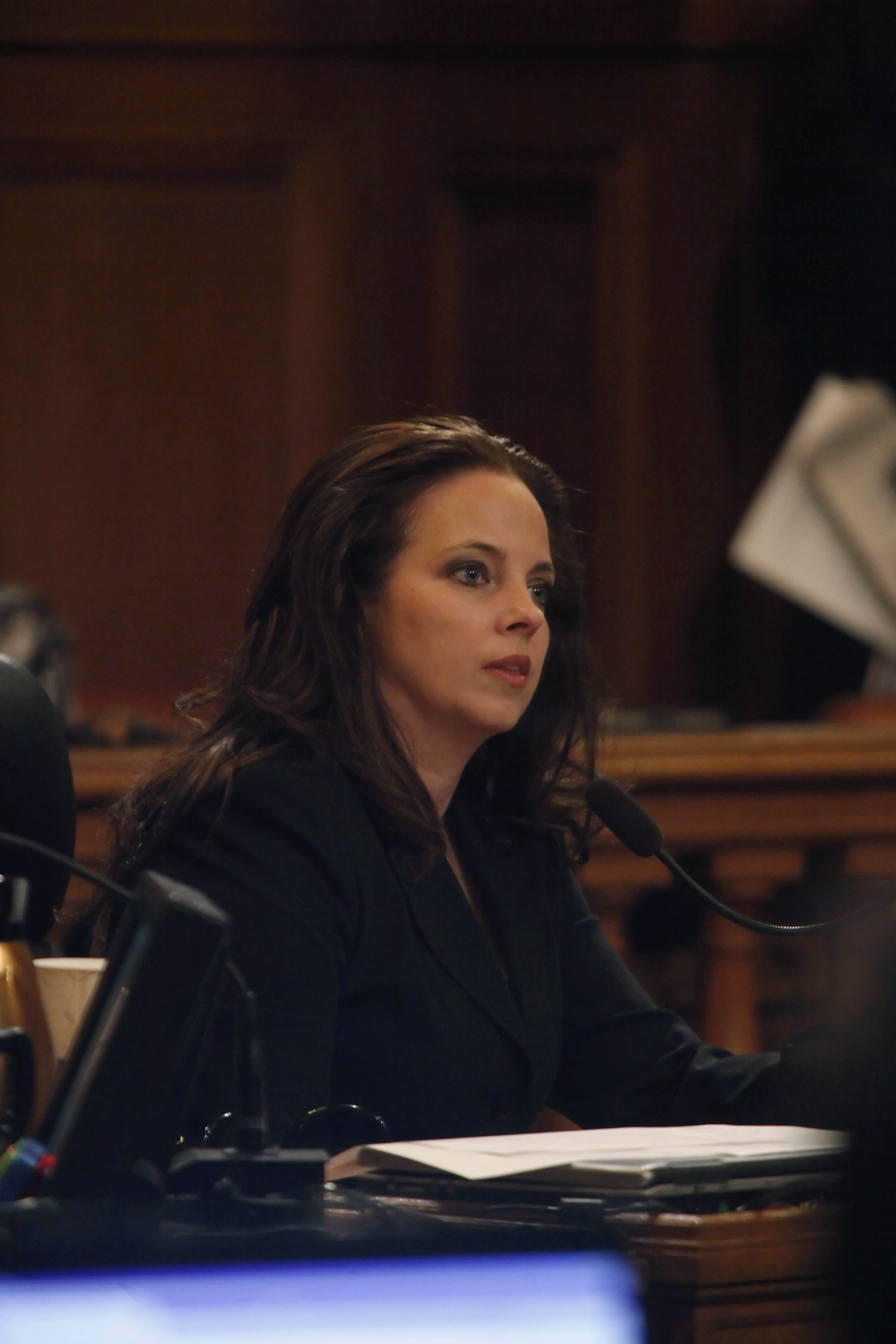 A woman in a black jacket sits at a desk with a microphone, papers in front of her, in a wood-paneled room.