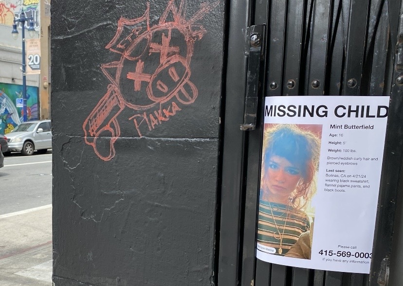 A graffiti of a gun with text and a "Missing Child" flyer featuring a young person is posted on an urban utility box.