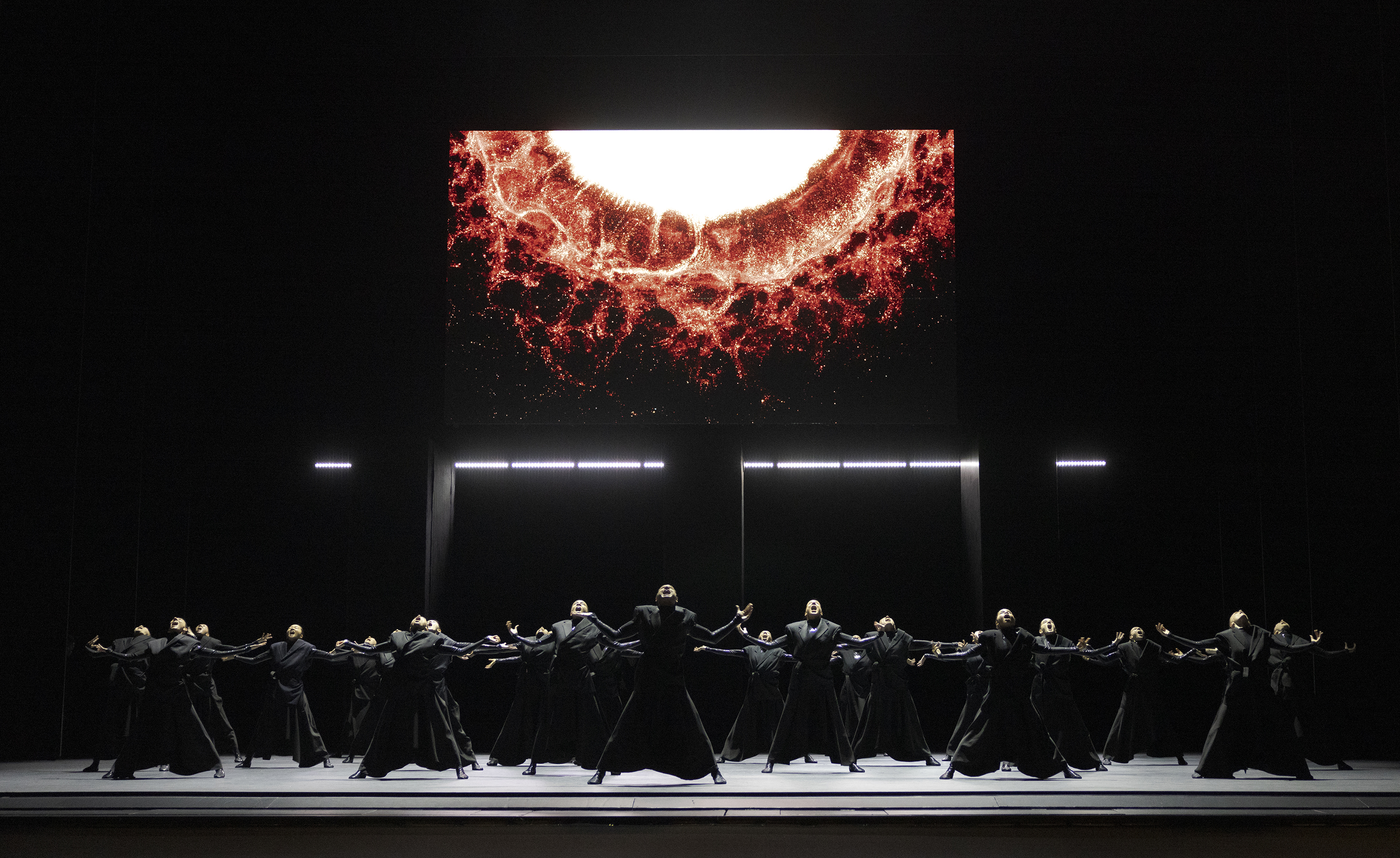 Dancers in black, outstretched, face a giant screen displaying a fiery cosmic image, all on a minimalist stage.