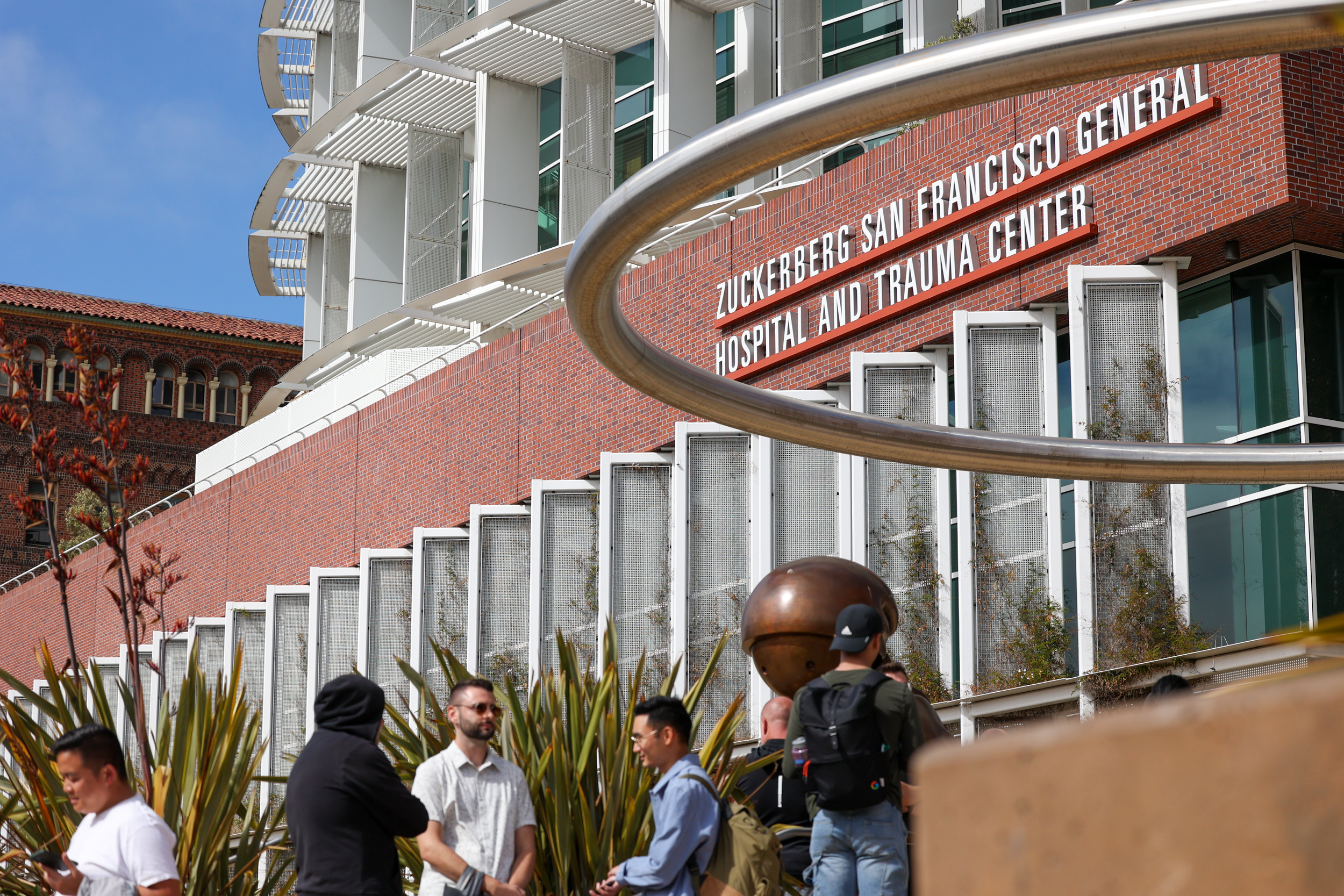 People converse outside a hospital with a large sign that reads &quot;Zuckerberg San Francisco General Hospital and Trauma Center&quot;.