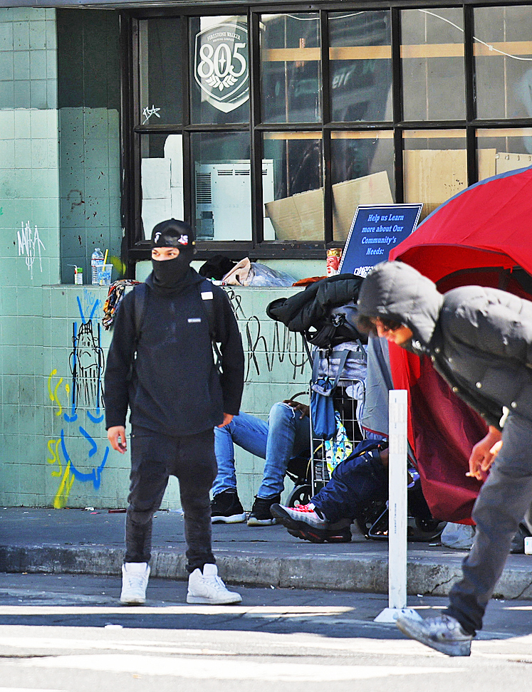 A person in a black mask and clothes stands on a sidewalk with graffiti and passing pedestrians near a storefront.