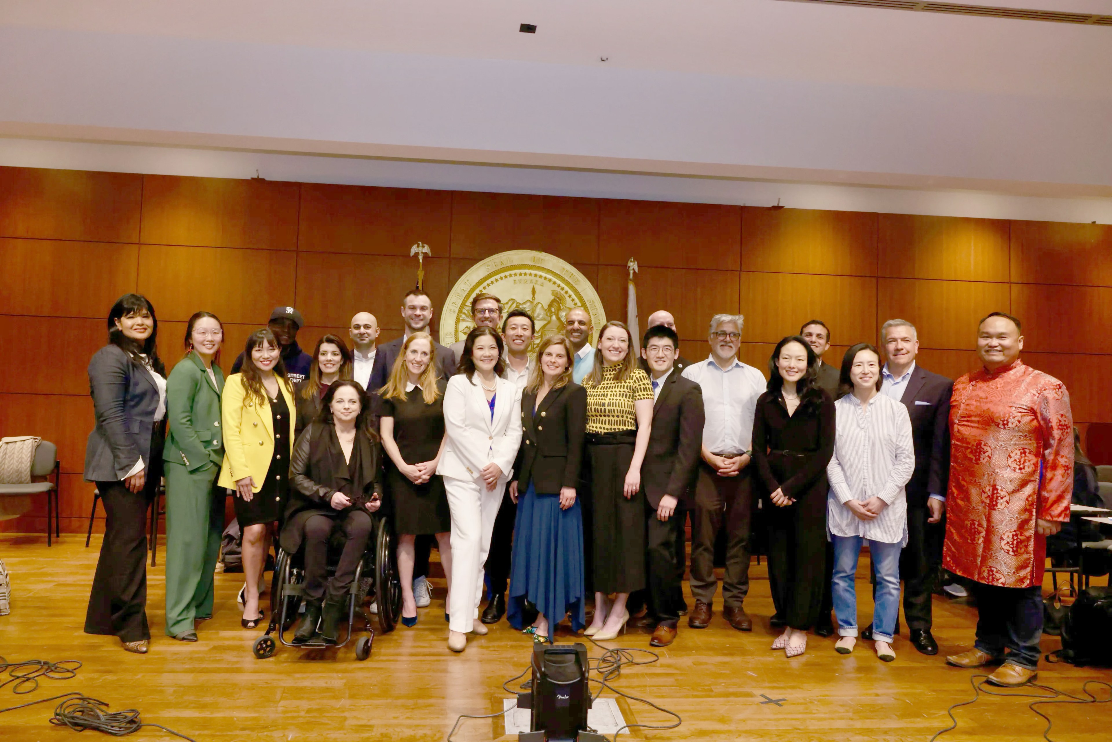 Group of diverse people posing in a room, some standing and one in a wheelchair, smiling at the camera.
