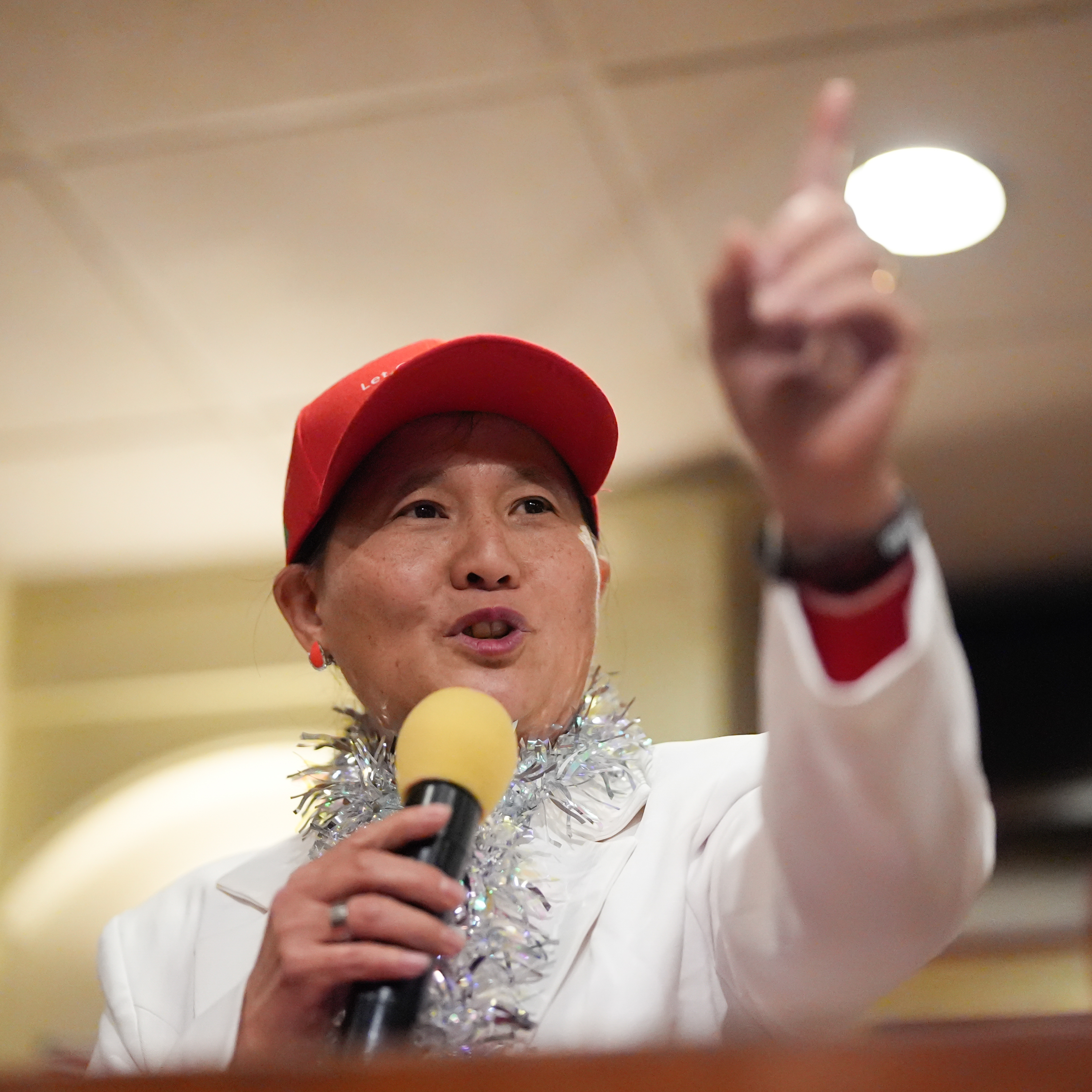 A person in a red cap and tinsel speaking into a microphone, gesturing upwards with one finger.