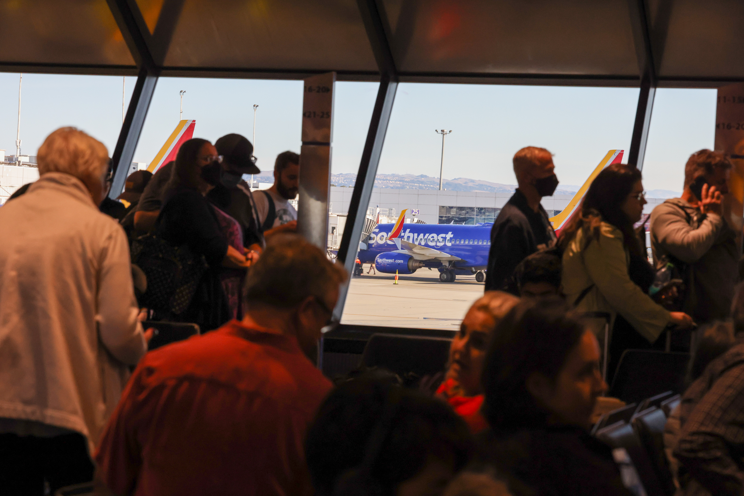Passengers in a waiting area with a blue airplane outside the window and mountains in the backdrop.