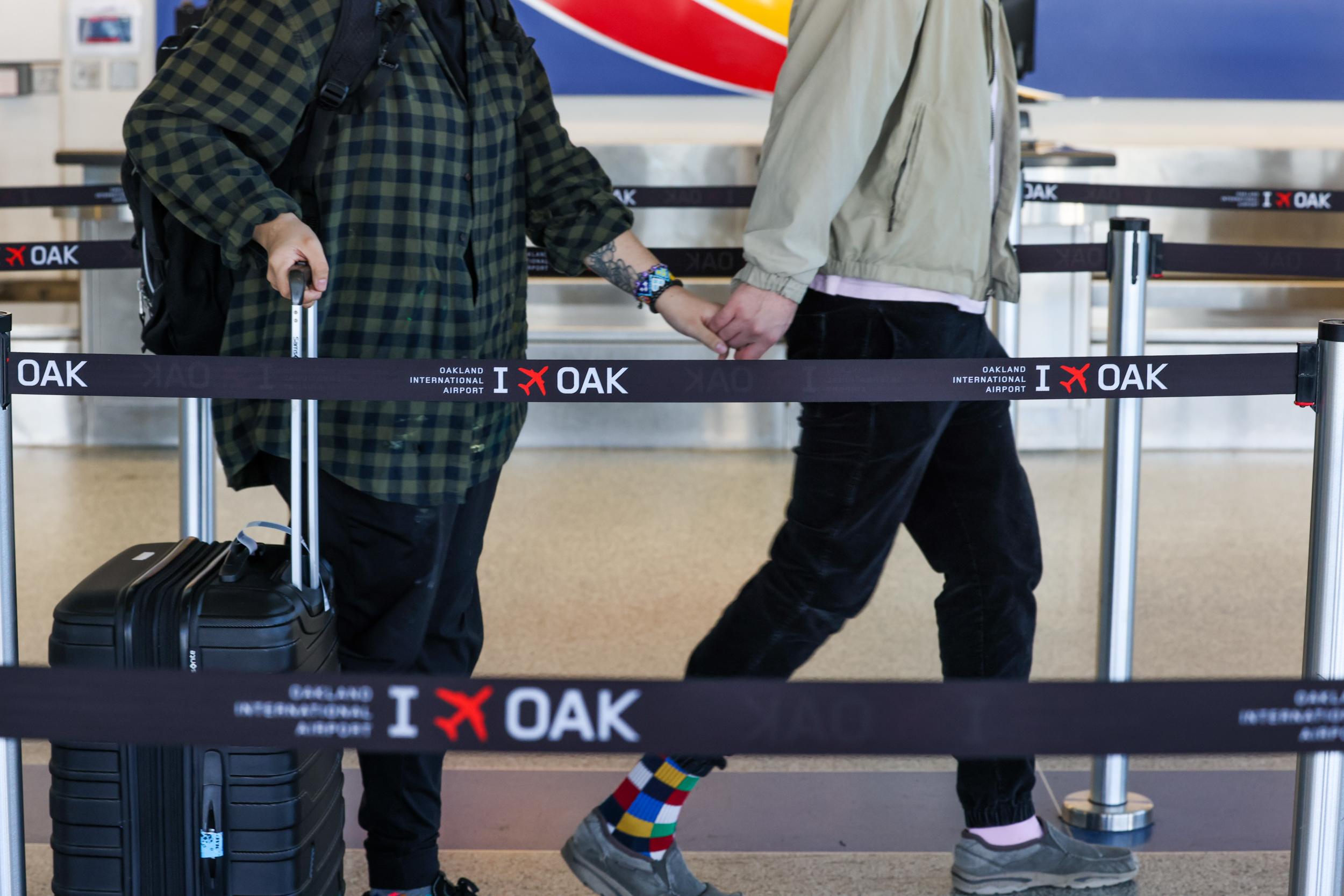 Two individuals at an airport hold hands behind a queue barrier, one with colorful socks, the other with a suitcase.