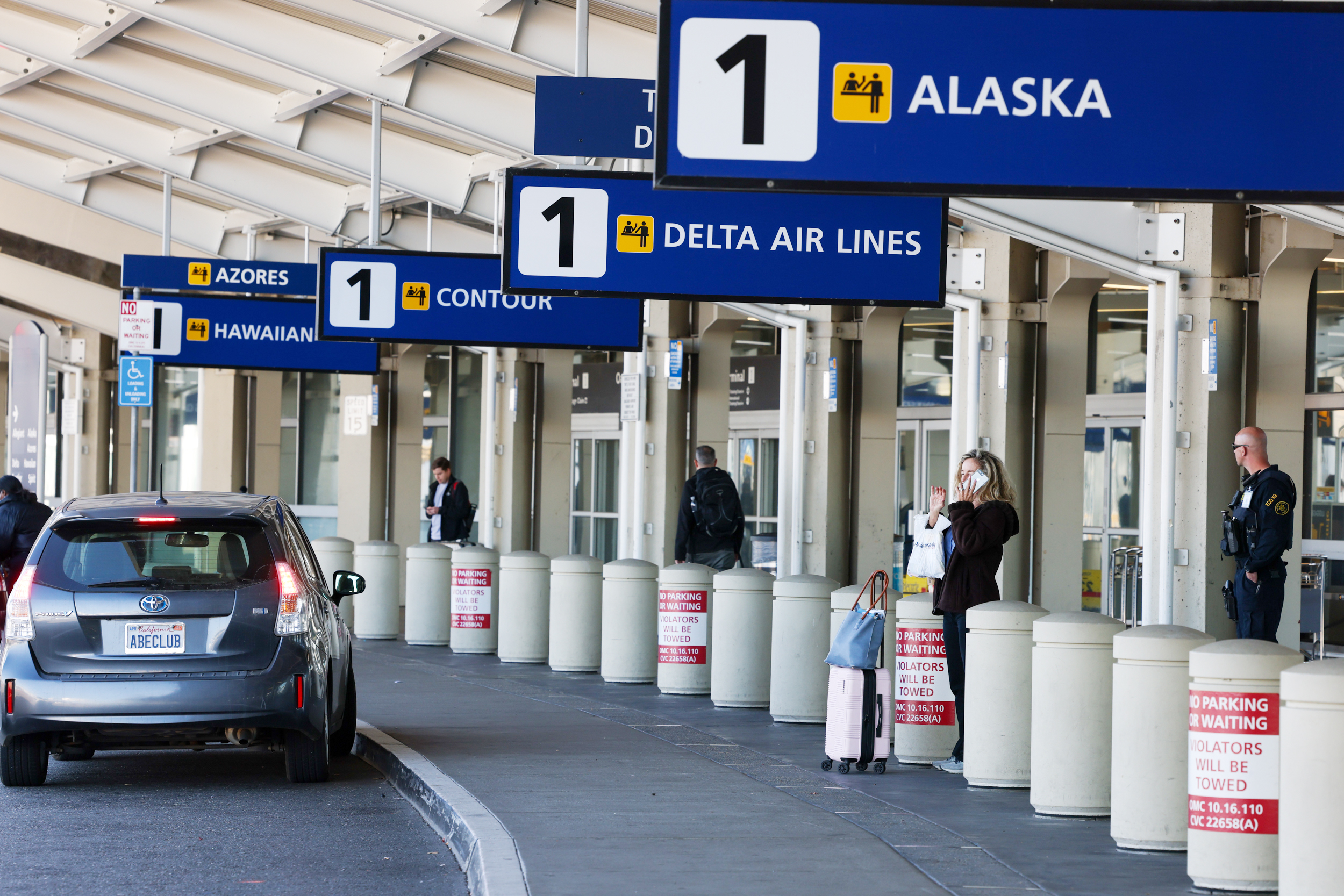 At an airport drop-off zone, people stand by signs for Alaska and Delta Air Lines; a car waits, and a security officer observes.