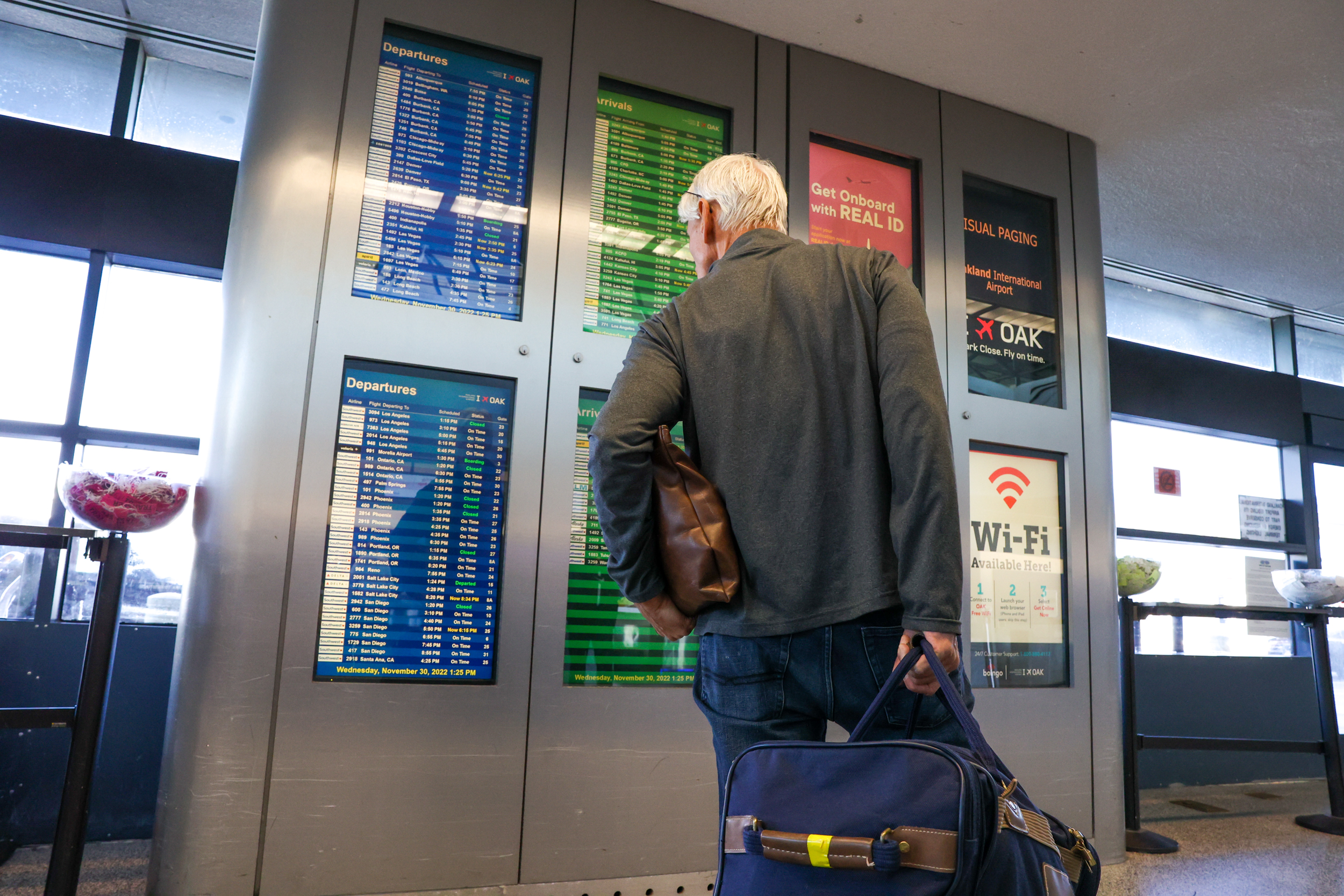 A person checks a flight information display board at an airport, carrying a shoulder bag. Signs for Wi-Fi and REAL ID are visible.
