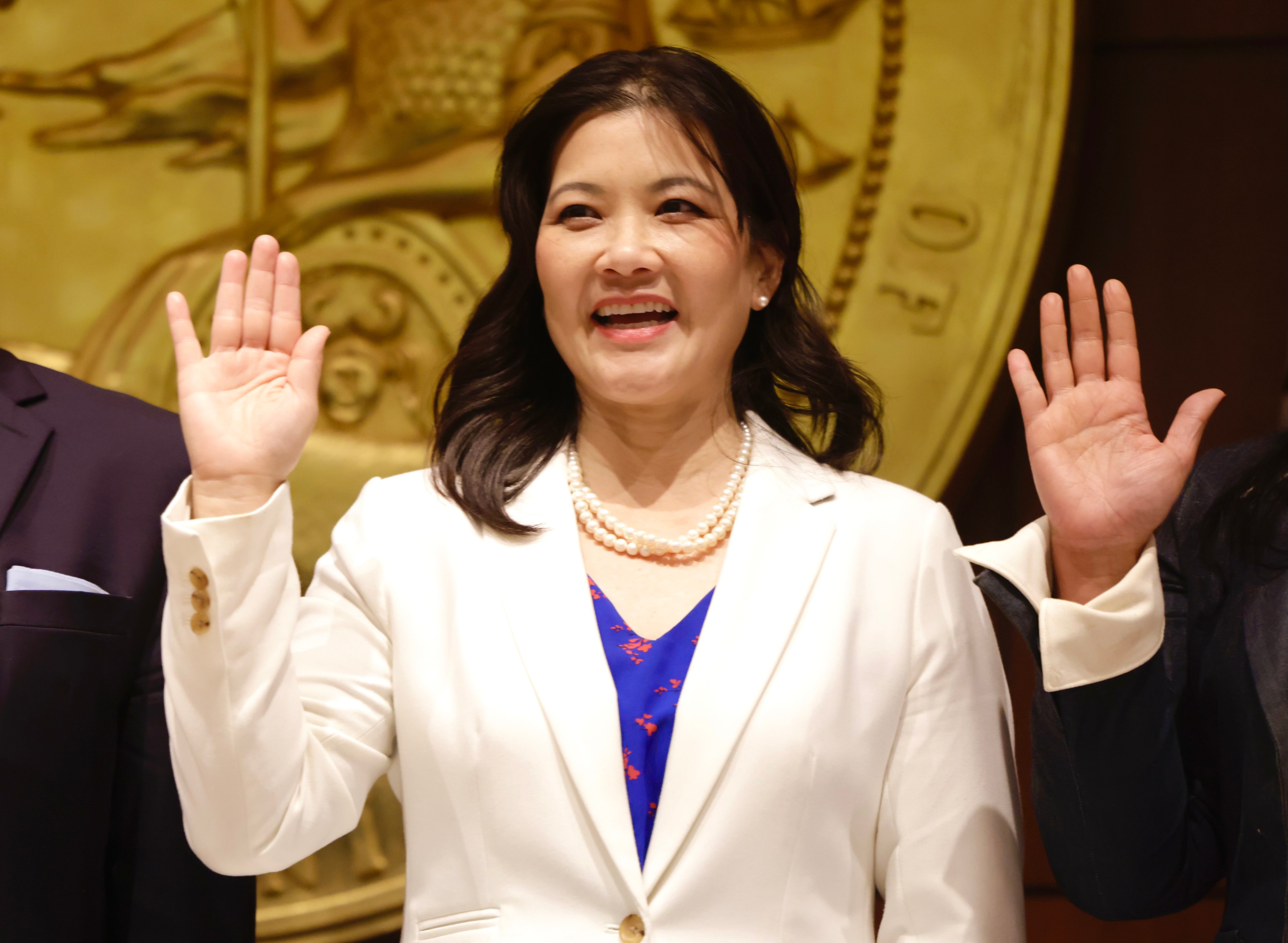 A woman in a white jacket is smiling and raising her hands, with ornate golden decor in the background.