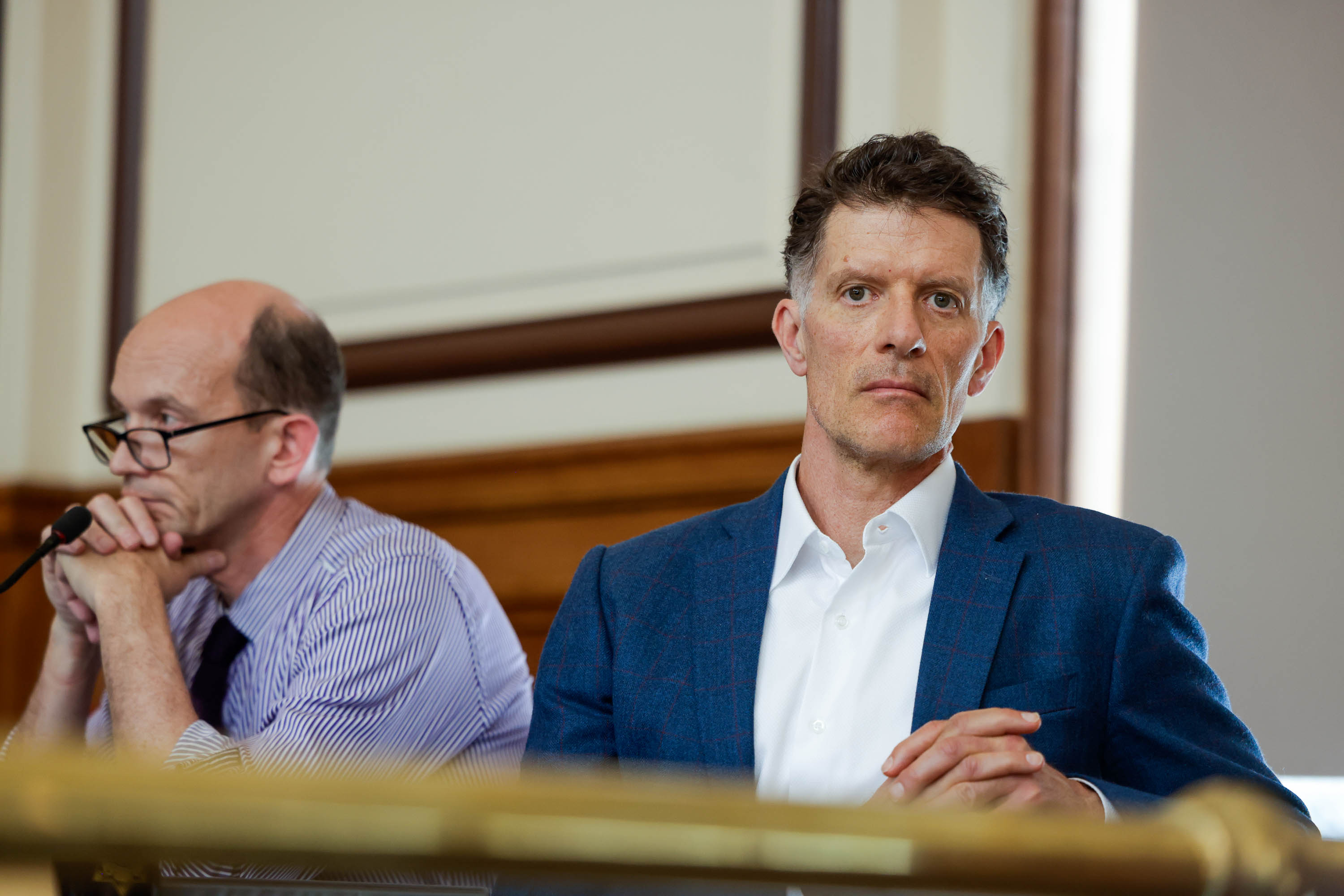 Two men sit in a room; one staring thoughtfully, the other with hands clasped, possibly in a meeting.