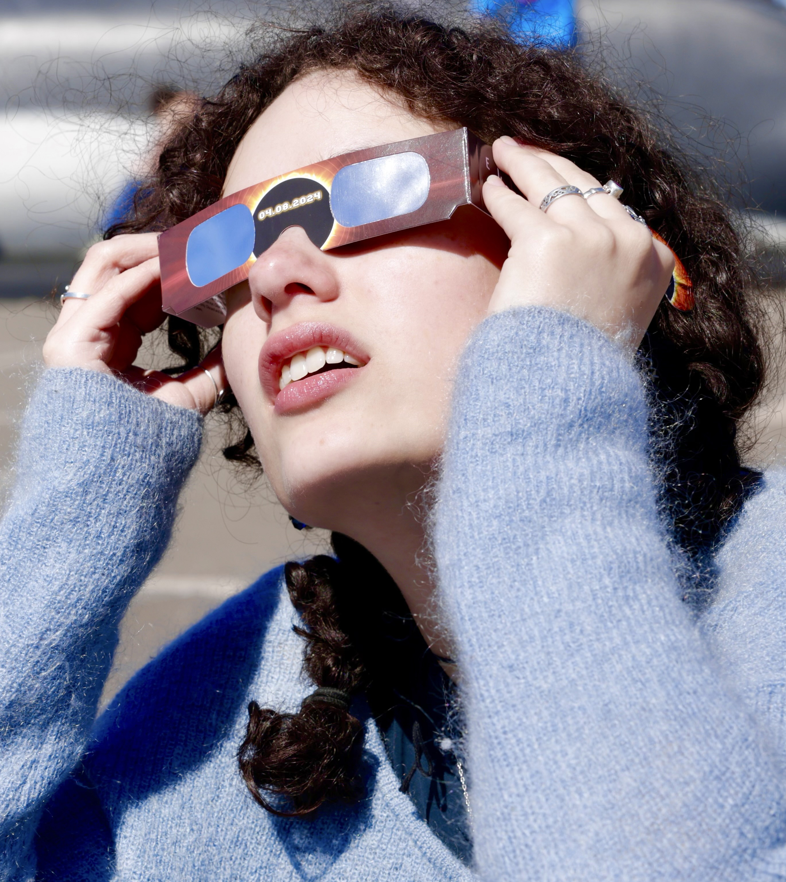 A person is wearing eclipse glasses and looking upwards, with curly hair and a blue sweater.
