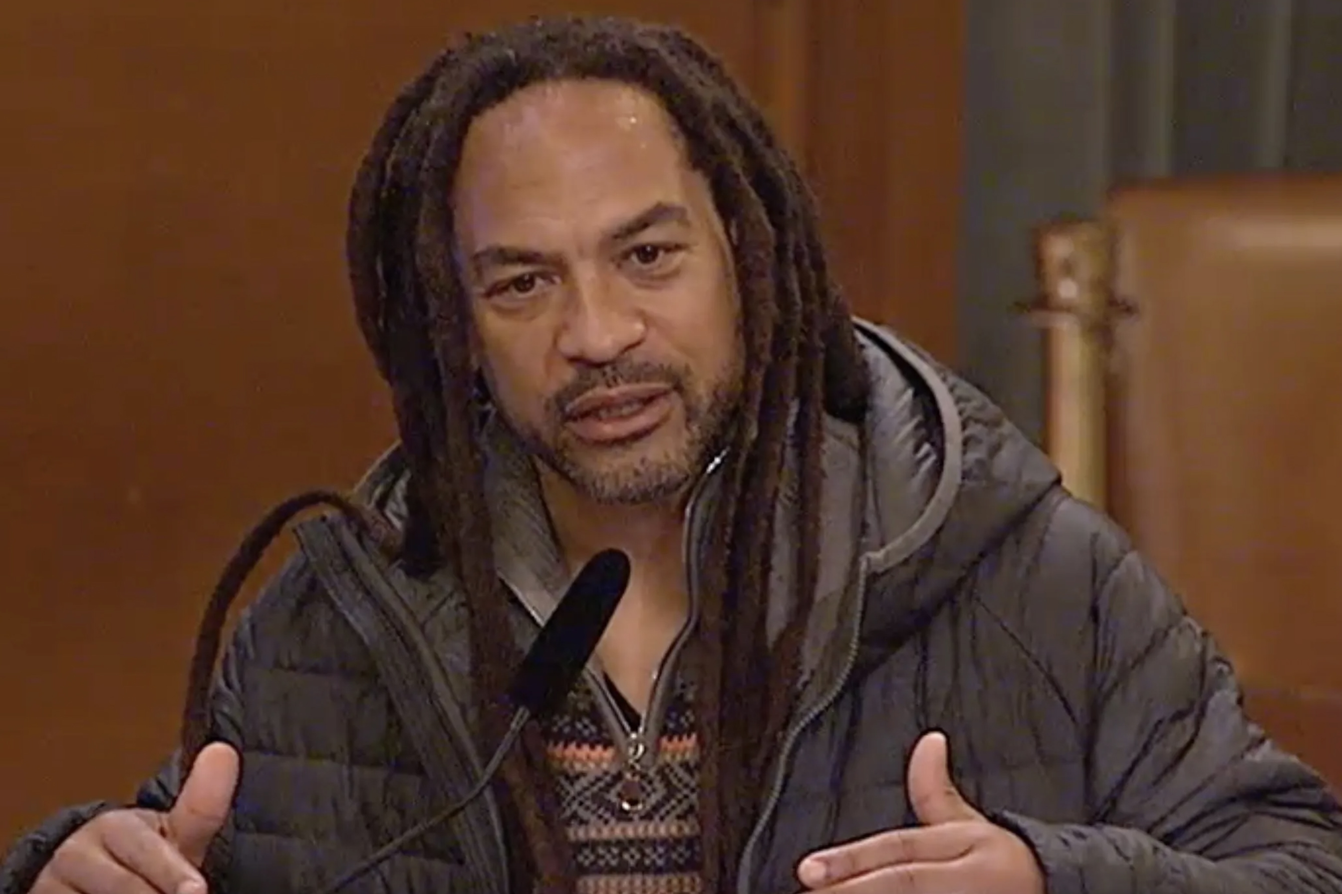 A man with dreadlocks wearing a puffy jacket is speaking into a microphone.