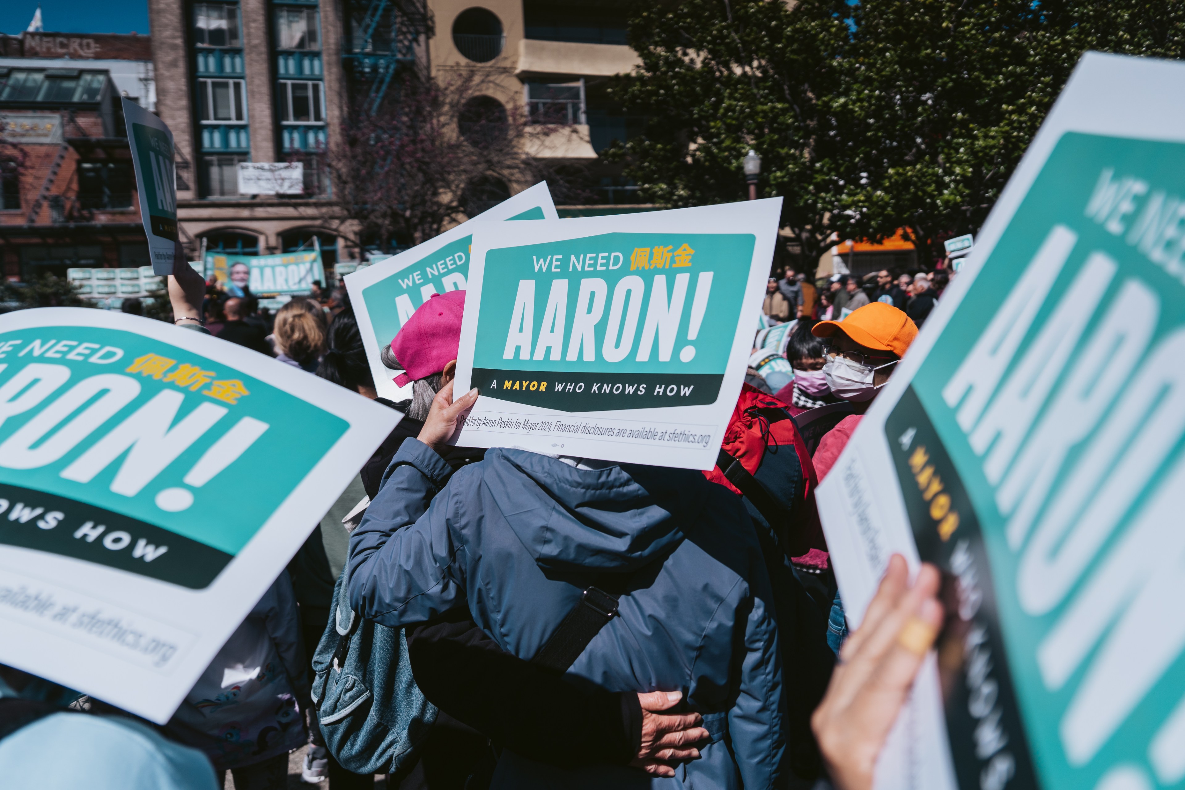 People at a rally hold signs supporting &quot;Aaron&quot; for mayor, with buildings in the background.