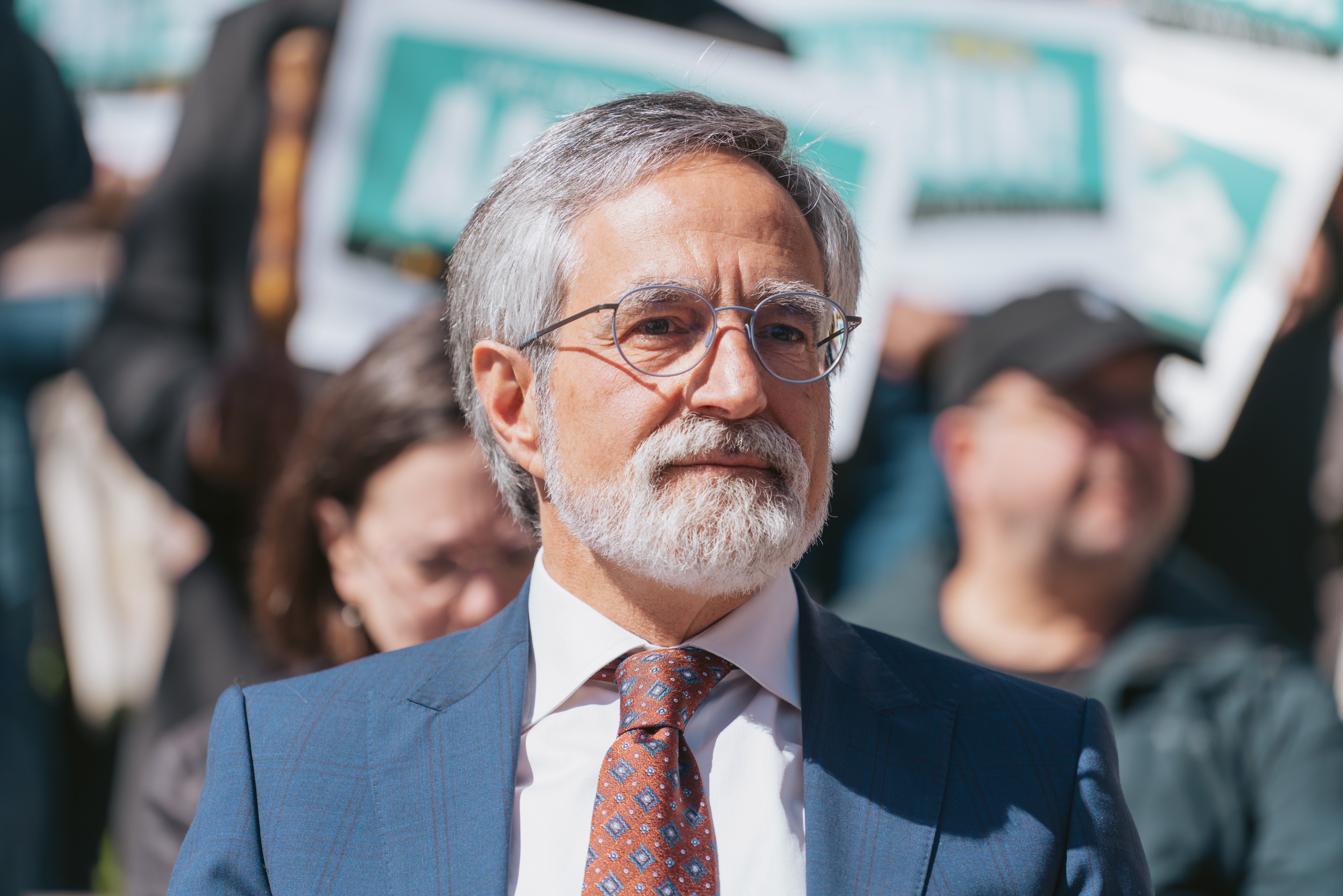 Supervisor Aaron Peskin wearing glasses and a suit is in focus at a daytime outdoor event, with blurry supporters and signs in the background.