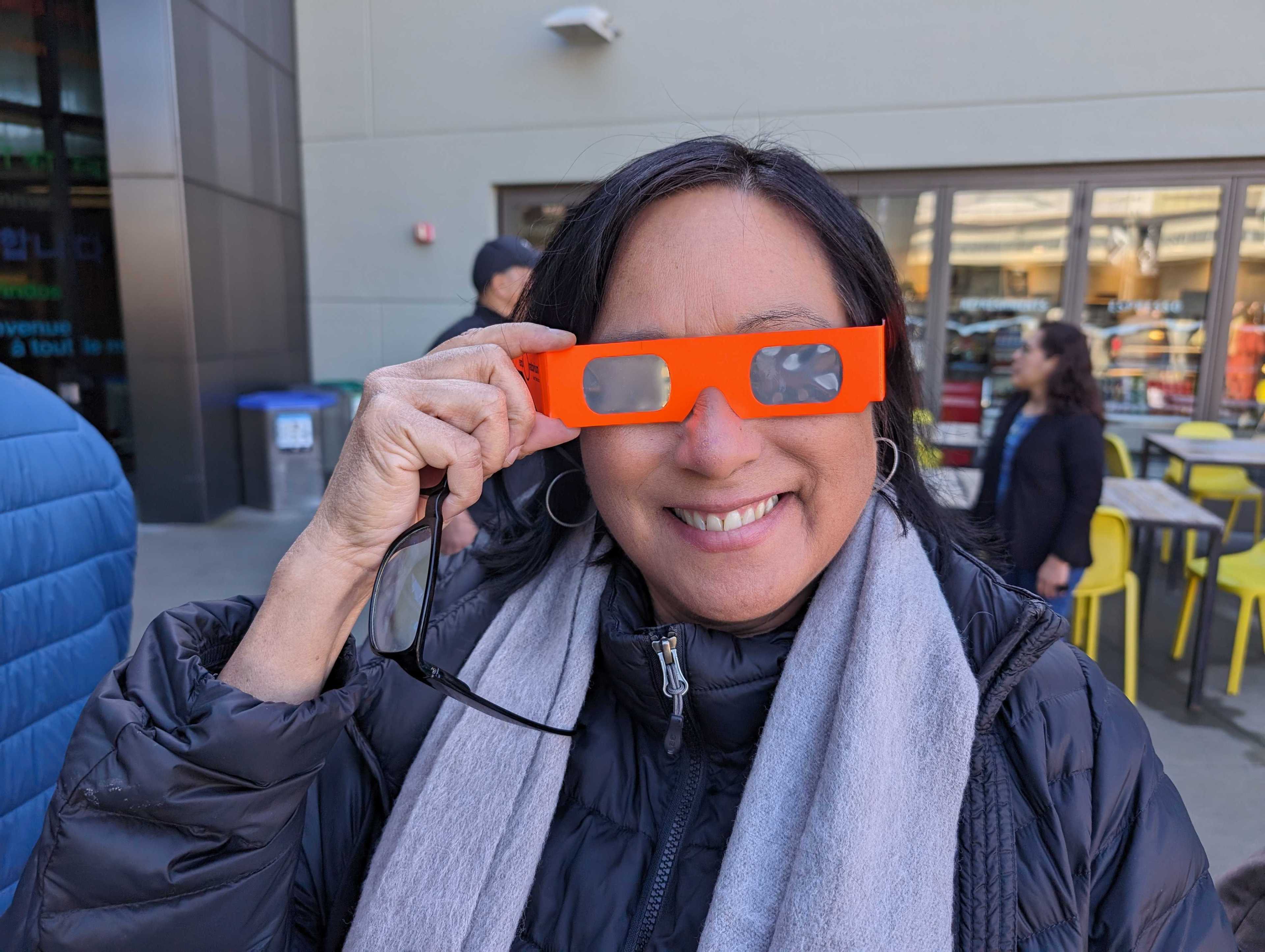 A smiling woman holds orange glasses with one rectangular lens missing.