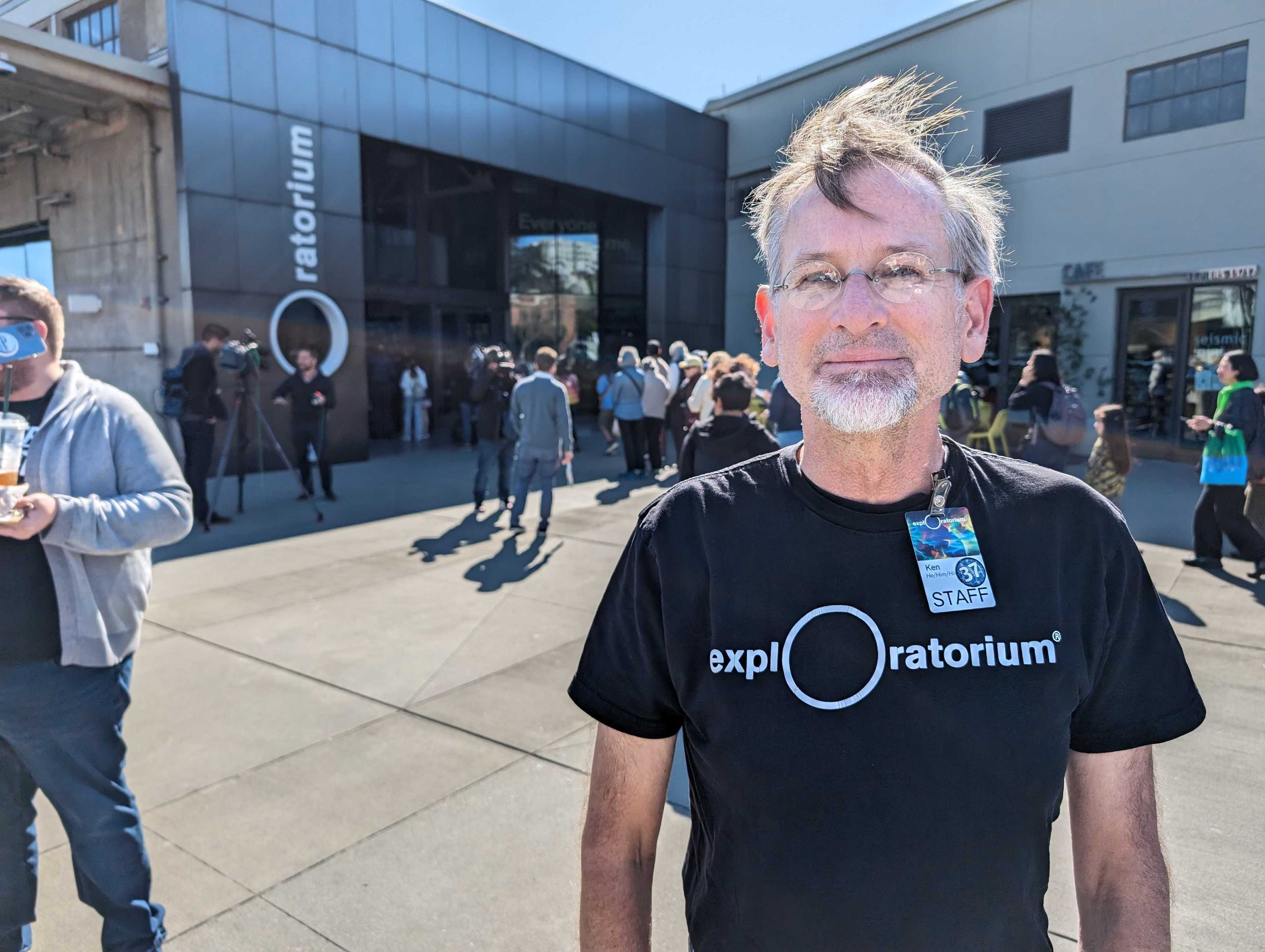 A man in a black Exploratorium staff shirt stands in front of a busy building entrance.