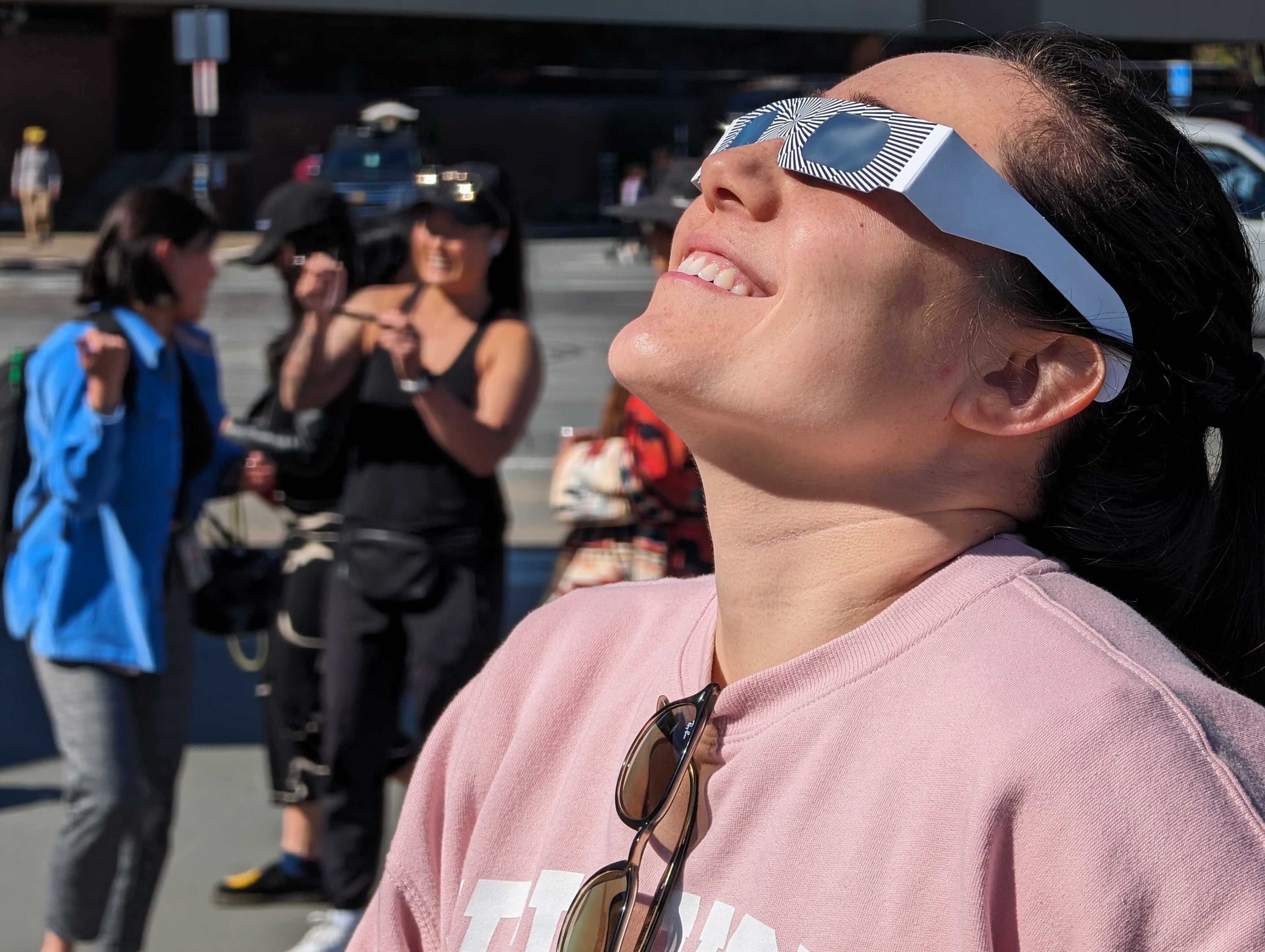 A smiling woman wearing unique sunglasses looks up, with blurred people in the background.