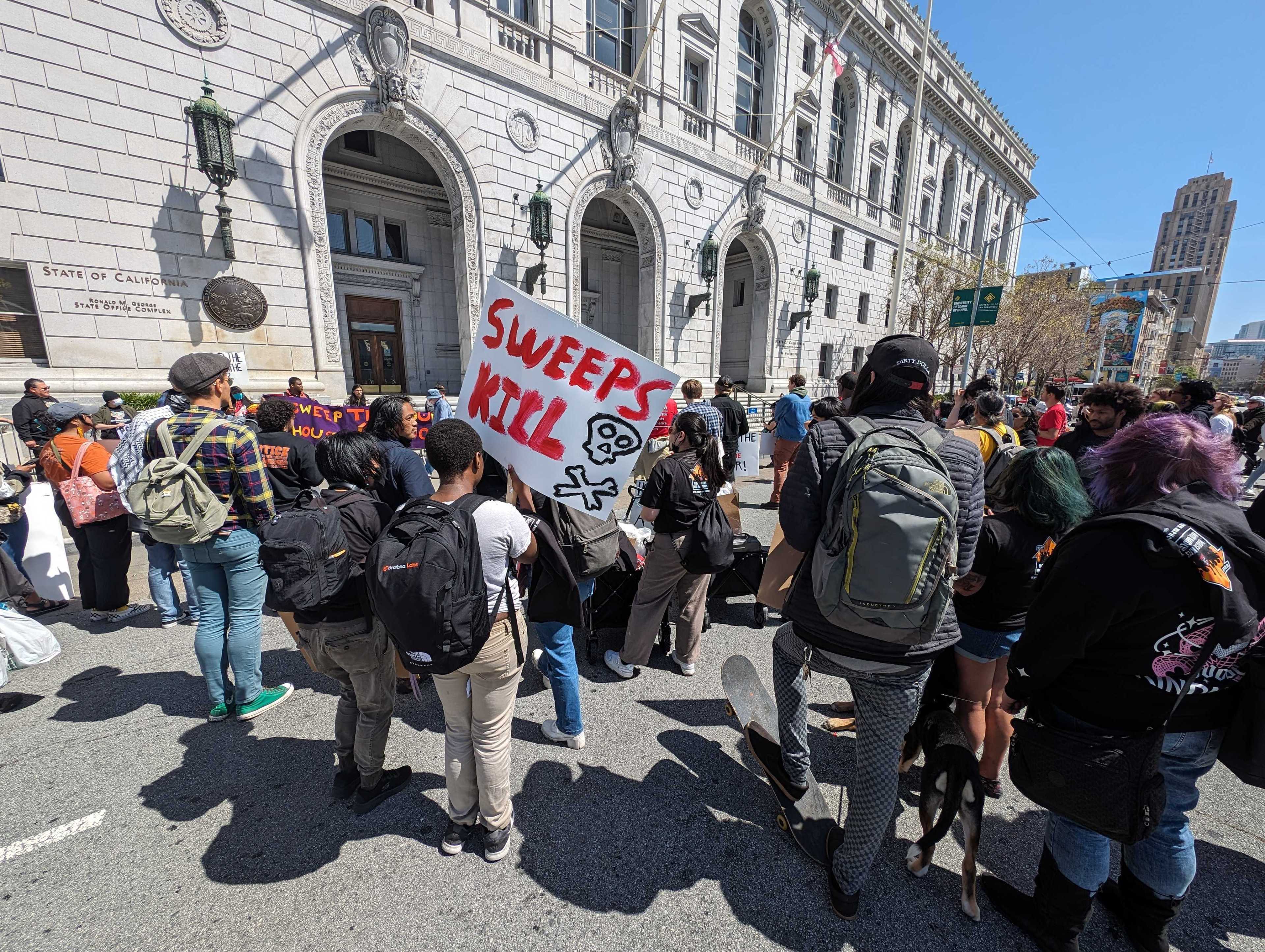 A group of protestors with signs, one reading "SWEEPS KILL", in front of a grand building on a sunny day.