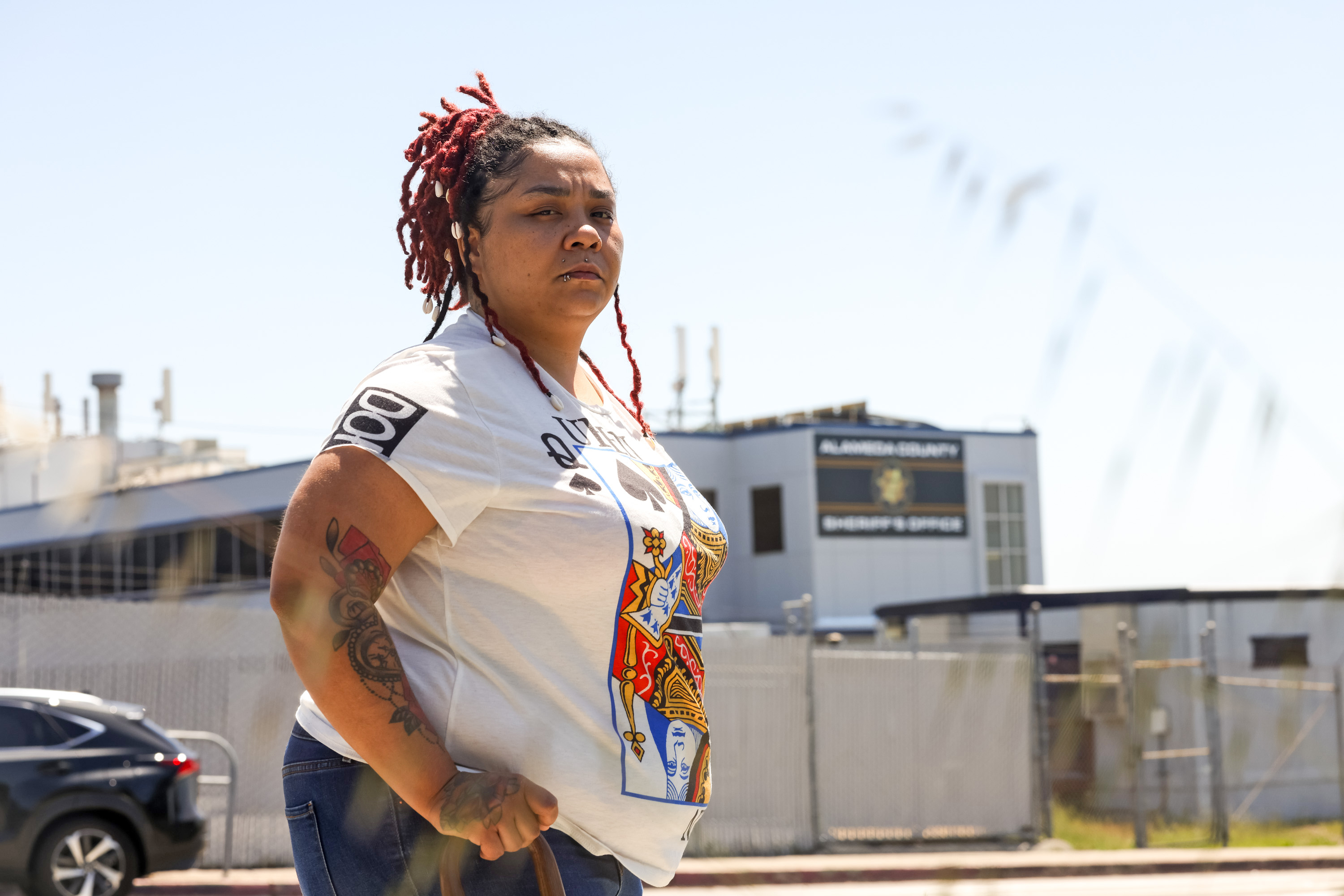 A person with red dreadlocks and tattoos stands confidently in front of an industrial backdrop, wearing a graphic t-shirt.