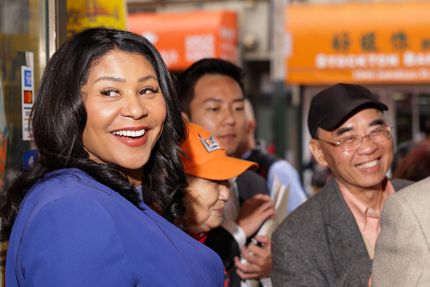 A smiling woman in a blue top is at the forefront with cheerful people in the background under orange signs with Asian characters.