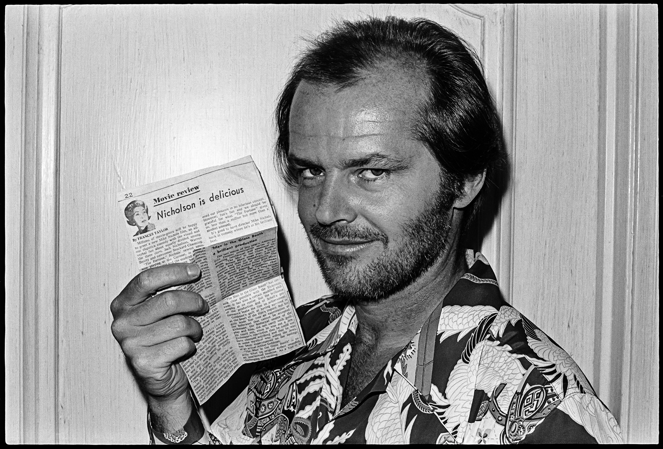 A man with a sly grin, wearing a printed shirt, holds a newspaper with a review titled "Nicholson is delicious."