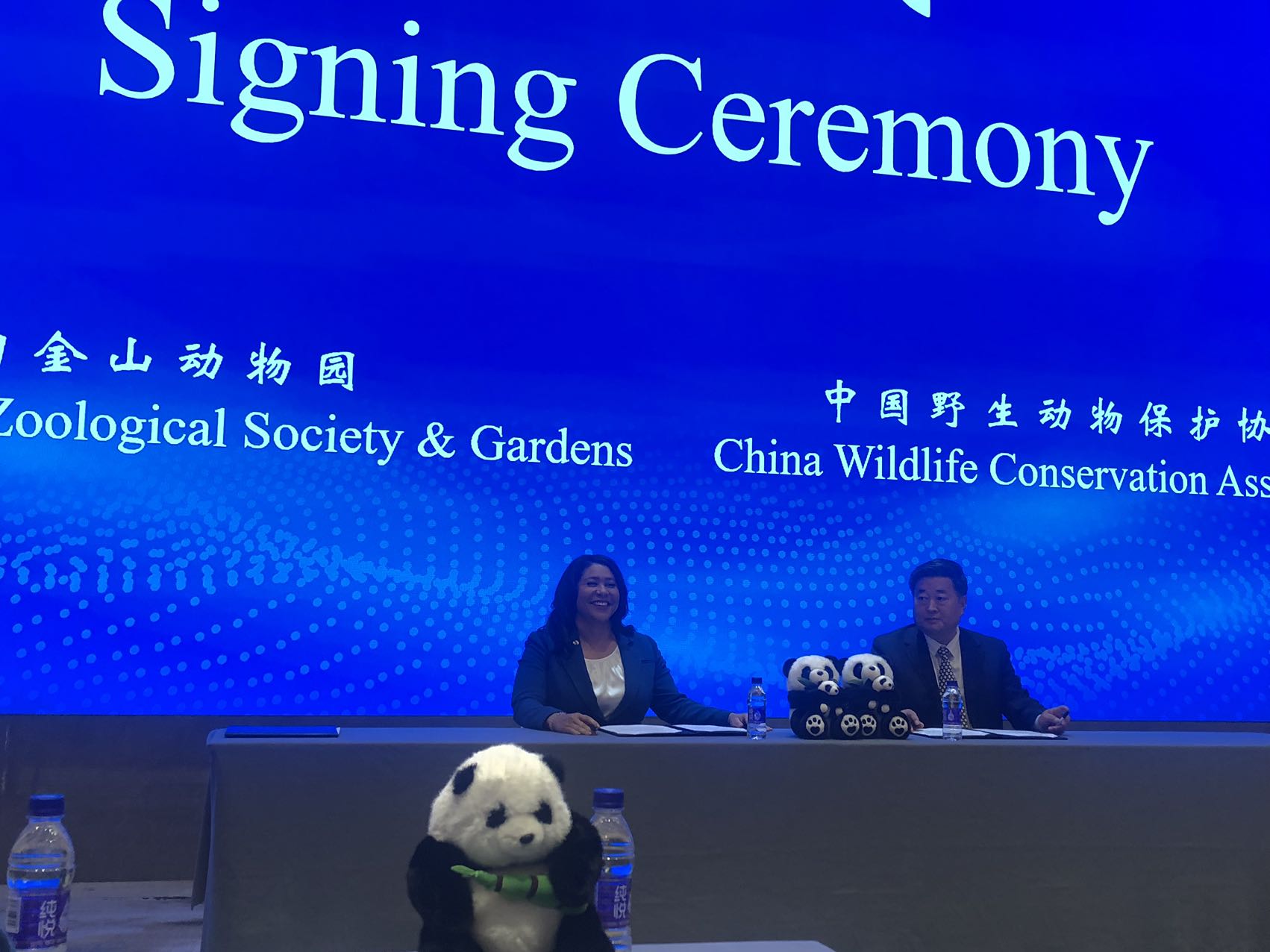 Two people are seated at a table with a &quot;Signing Ceremony&quot; backdrop, flanked by plush panda toys.