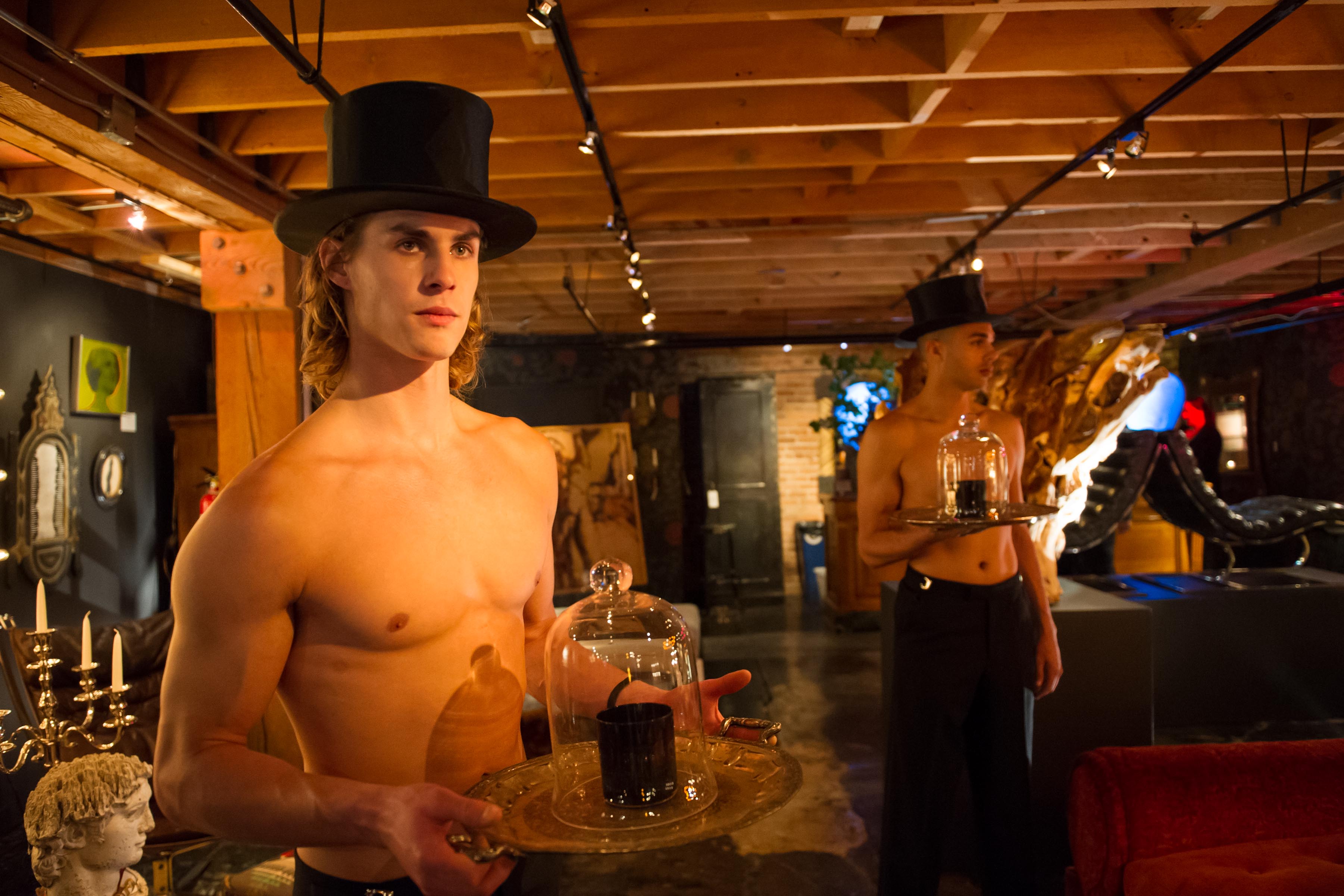 Two shirtless men wearing top hats hold trays with candles covered by glass domes in a dimly lit, eclectic room adorned with art and decorative items.