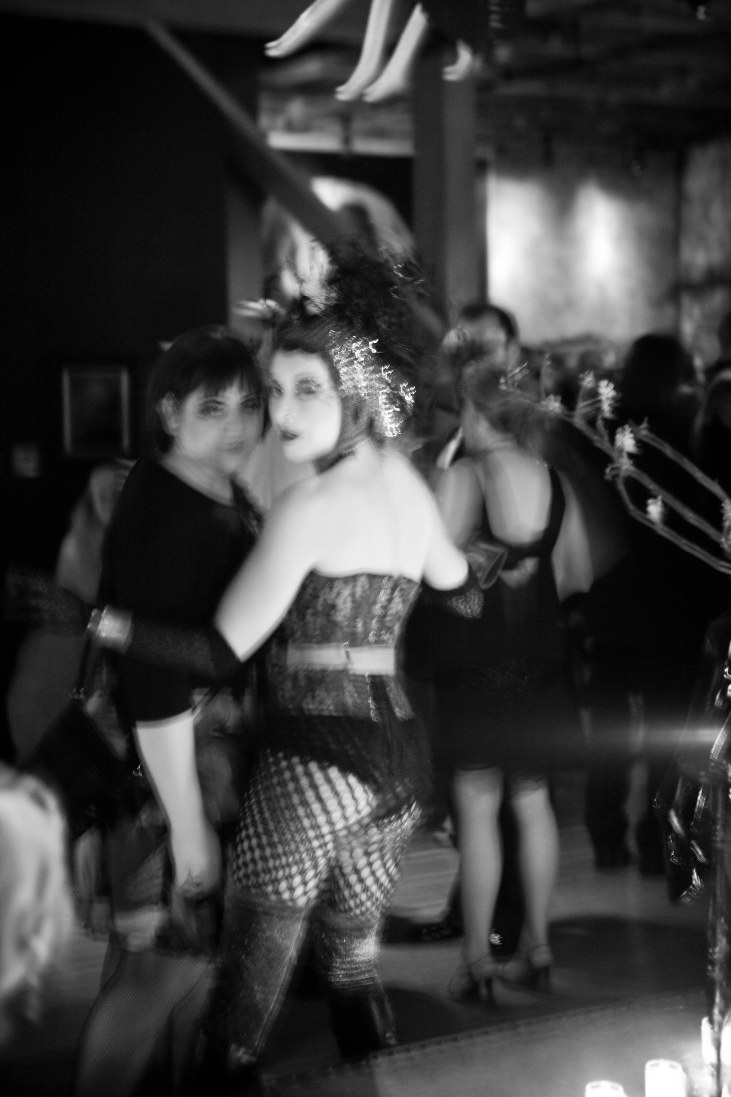 A black and white image shows two people in elaborate costumes, one with a feathered headpiece. Blurry effects and multiple other figures are in the dimly lit background.