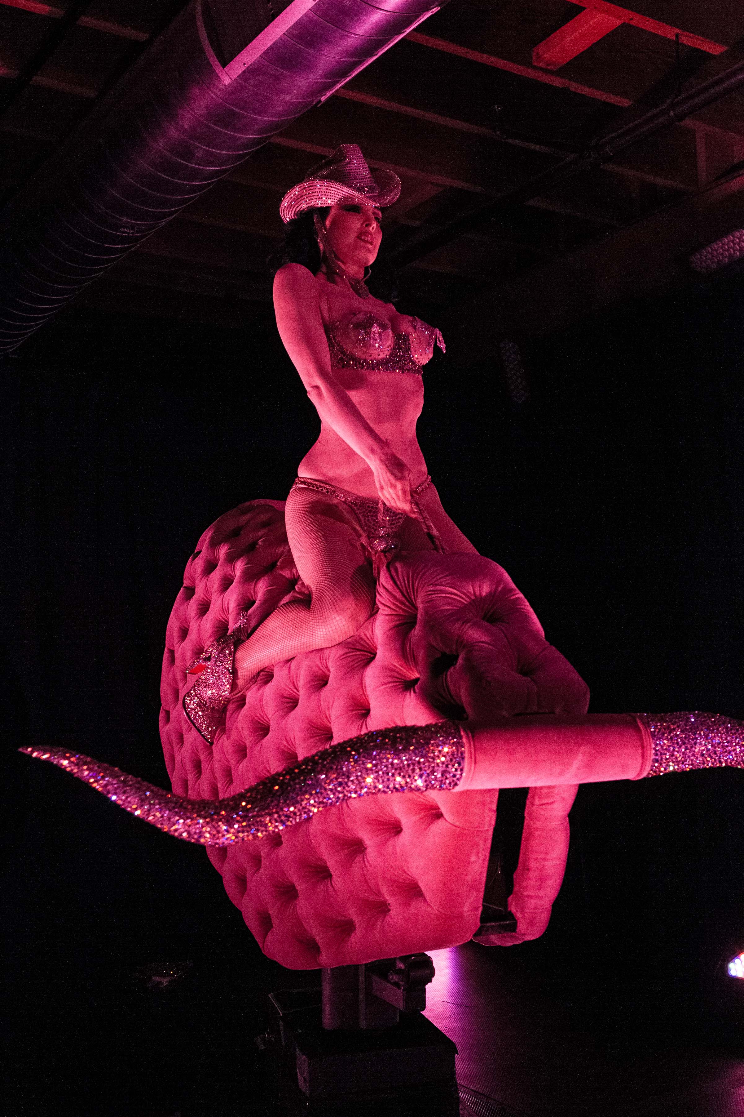 A woman in sparkly attire and a cowboy hat rides a pink upholstered mechanical bull with glittering horns, set in a dimly lit environment with purple tones.