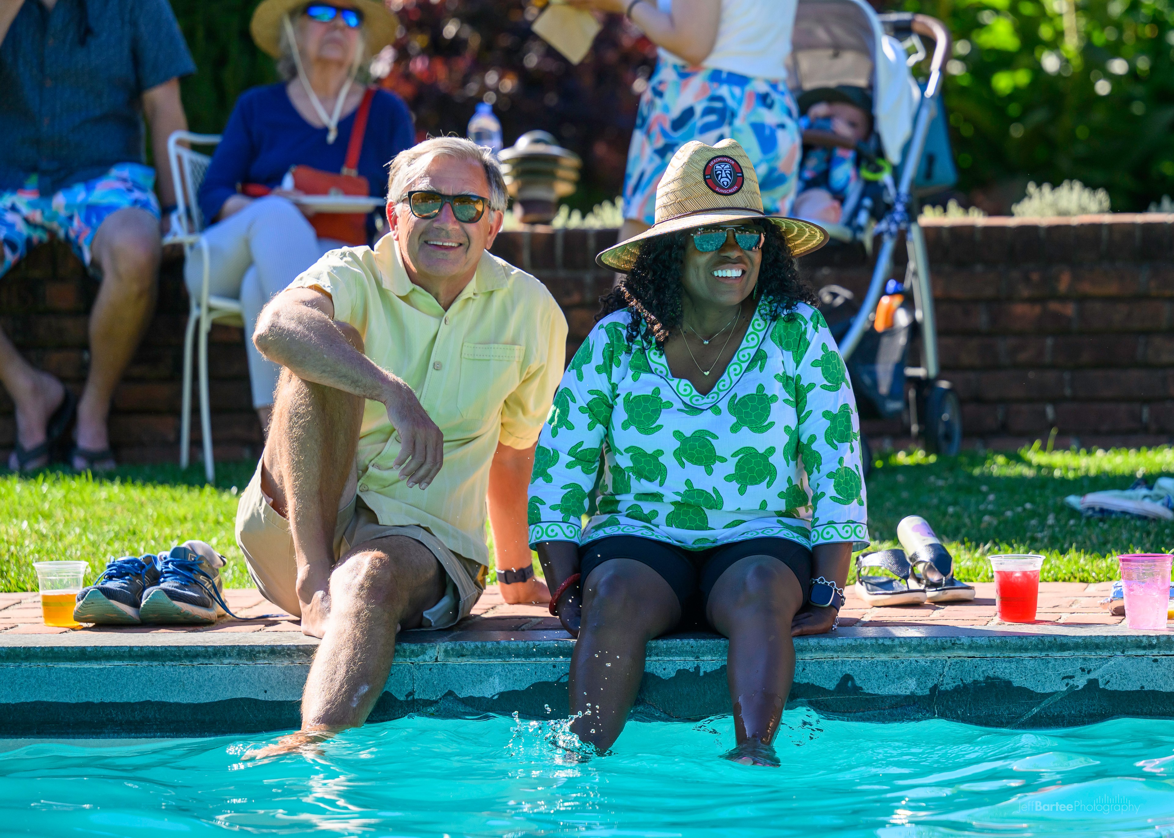Two people sit by a pool, smiling and dipping their feet into the water. They are wearing summer clothes and hats, with drinks and belongings beside them.